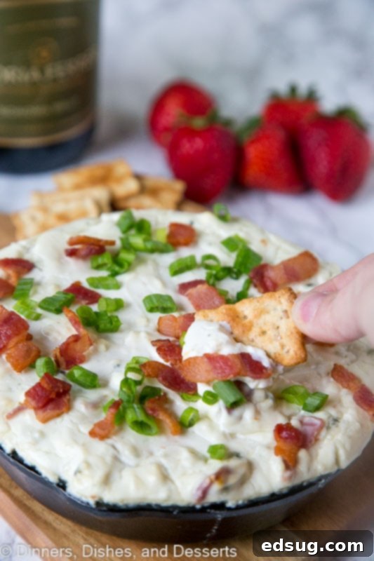 A close-up of baked blue cheese dip with green onions and bacon, served with a cracker ready for dipping
