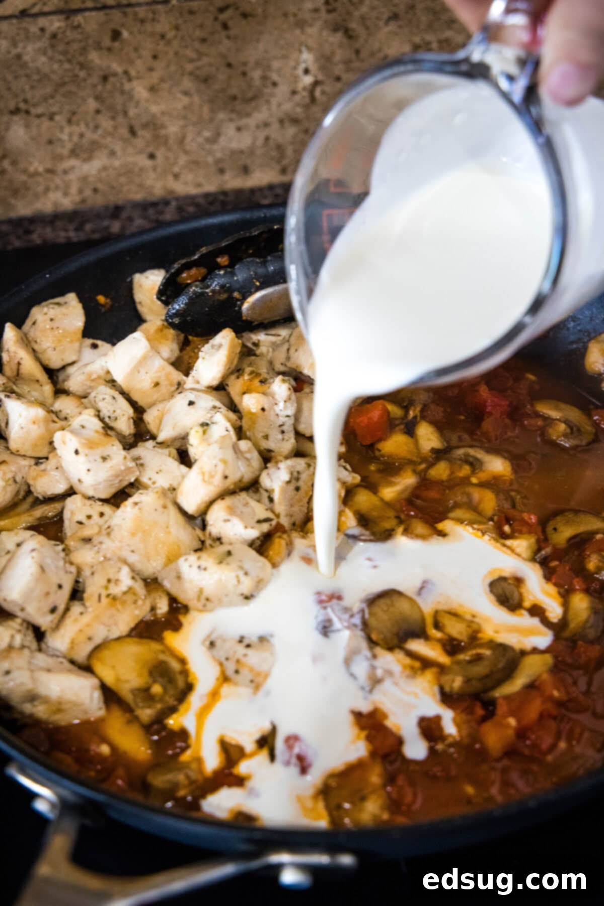 Creamy Tuscan Chicken Pasta 4 A hand pouring heavy cream into a large skillet containing sautéed chicken and diced tomatoes, creating a creamy sauce.
