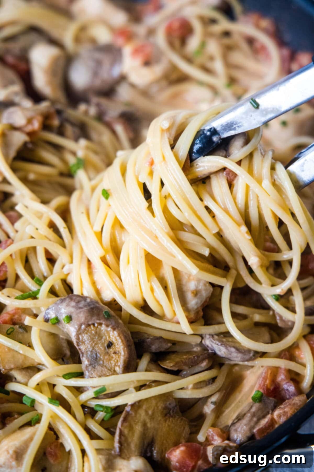Creamy Tuscan Chicken Pasta 6 Tongs lifting a generous portion of creamy Tuscan chicken pasta from a skillet, showing the well-coated pasta and pieces of chicken and mushrooms.