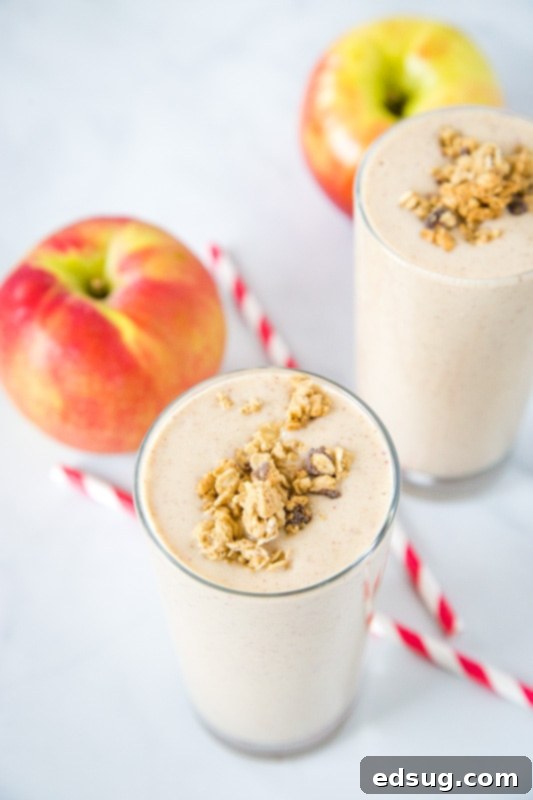 A vibrant apple cinnamon smoothie in a glass with a whole, fresh red apple and a straw beside it, emphasizing natural ingredients.