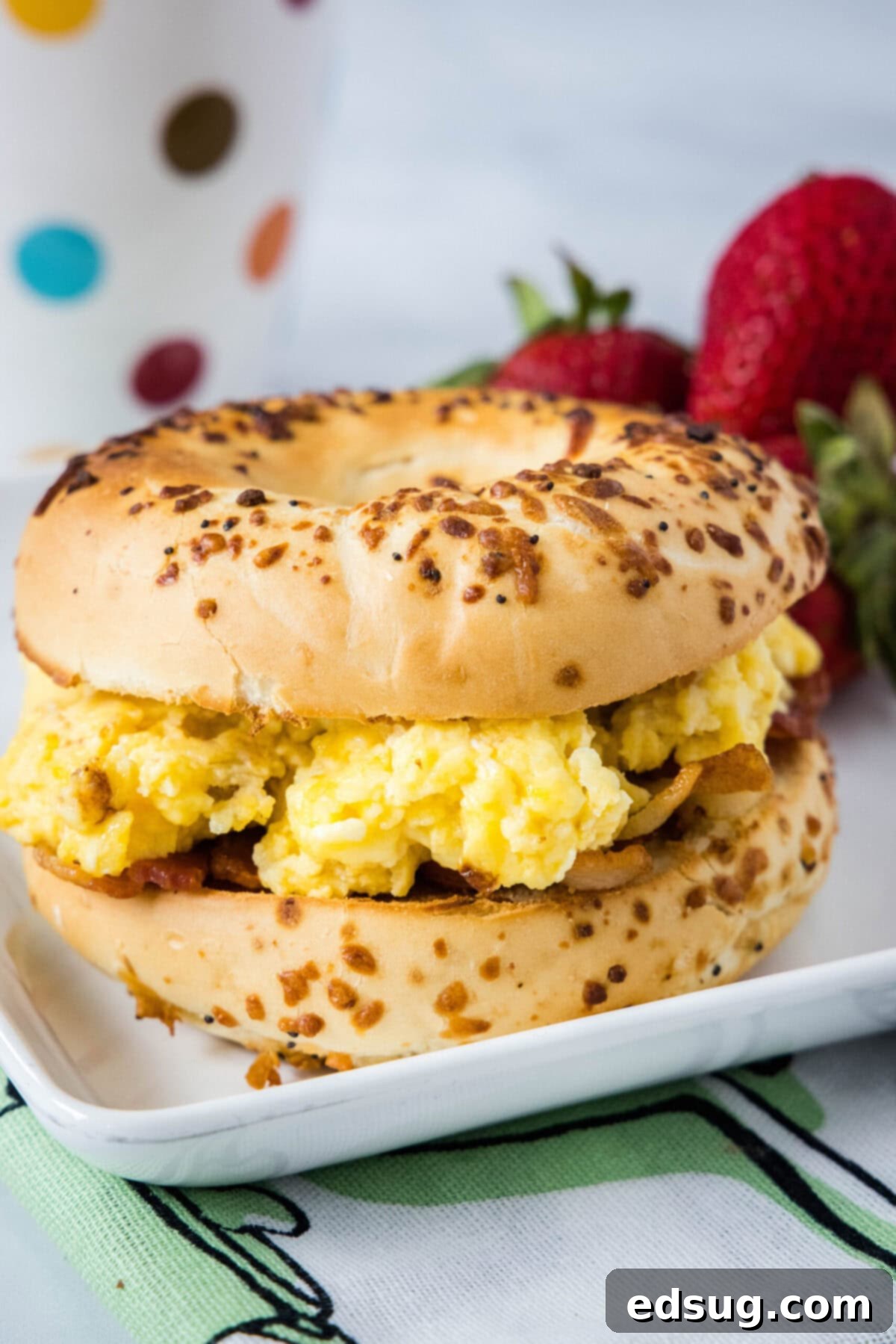This classic bacon egg and cheese sandwich is the ultimate breakfast, with fluffy eggs, melty cheese, crispy bacon, and a toasted bagel. A bagel sandwich with eggs on a plate with strawberries