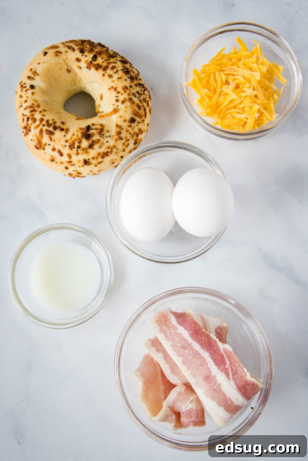 This classic bacon egg and cheese sandwich is the ultimate breakfast, with fluffy eggs, melty cheese, crispy bacon, and a toasted bagel. Overhead view of the ingredients needed for a bacon egg and cheese sandwich: a bagel, a bowl of bacon, a bowl of shredded cheese, a bowl of milk, and two eggs