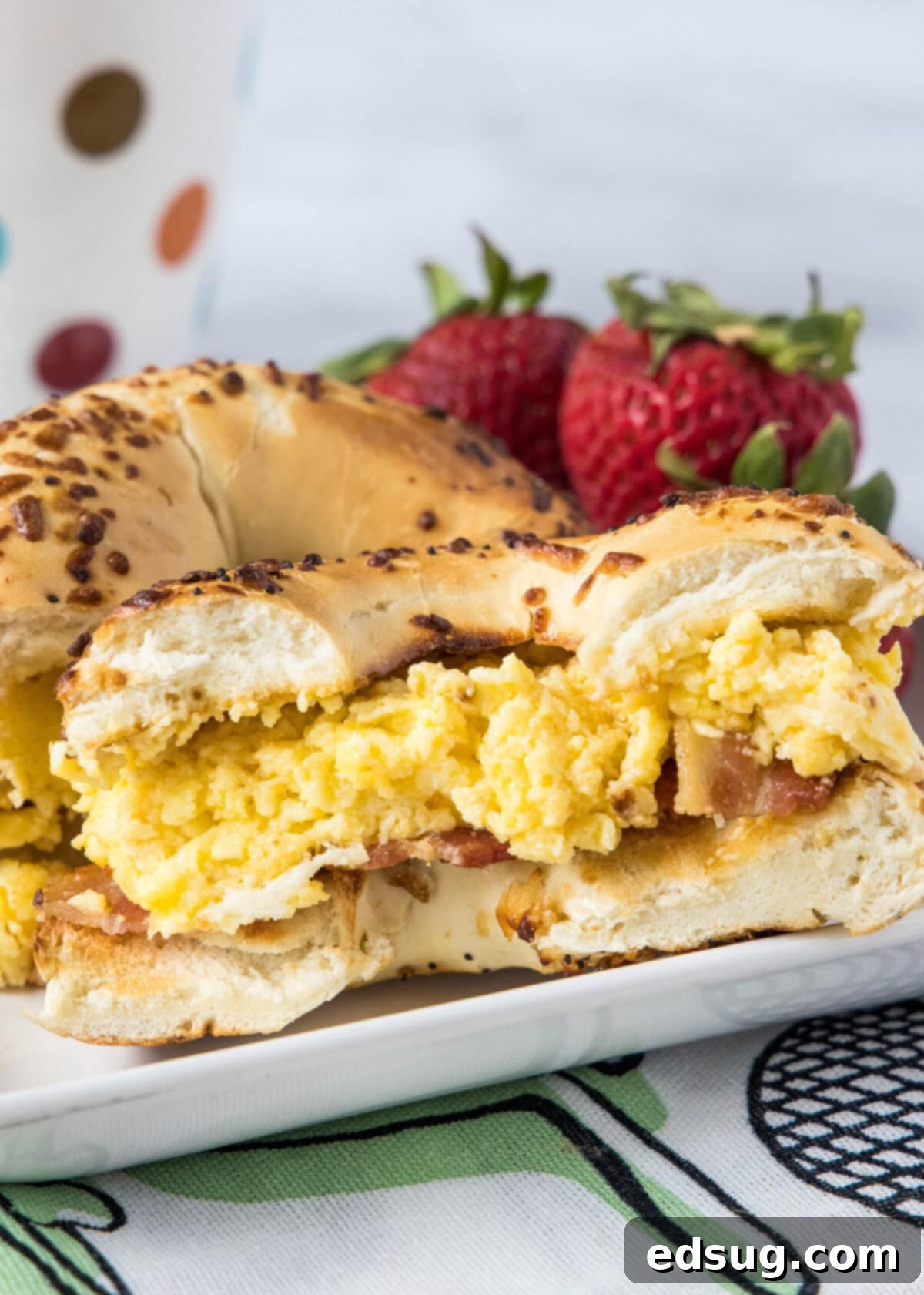 This classic bacon egg and cheese sandwich is the ultimate breakfast, with fluffy eggs, melty cheese, crispy bacon, and a toasted bagel. A bacon and egg breakfast sandwich cut in half on a plate with strawberries