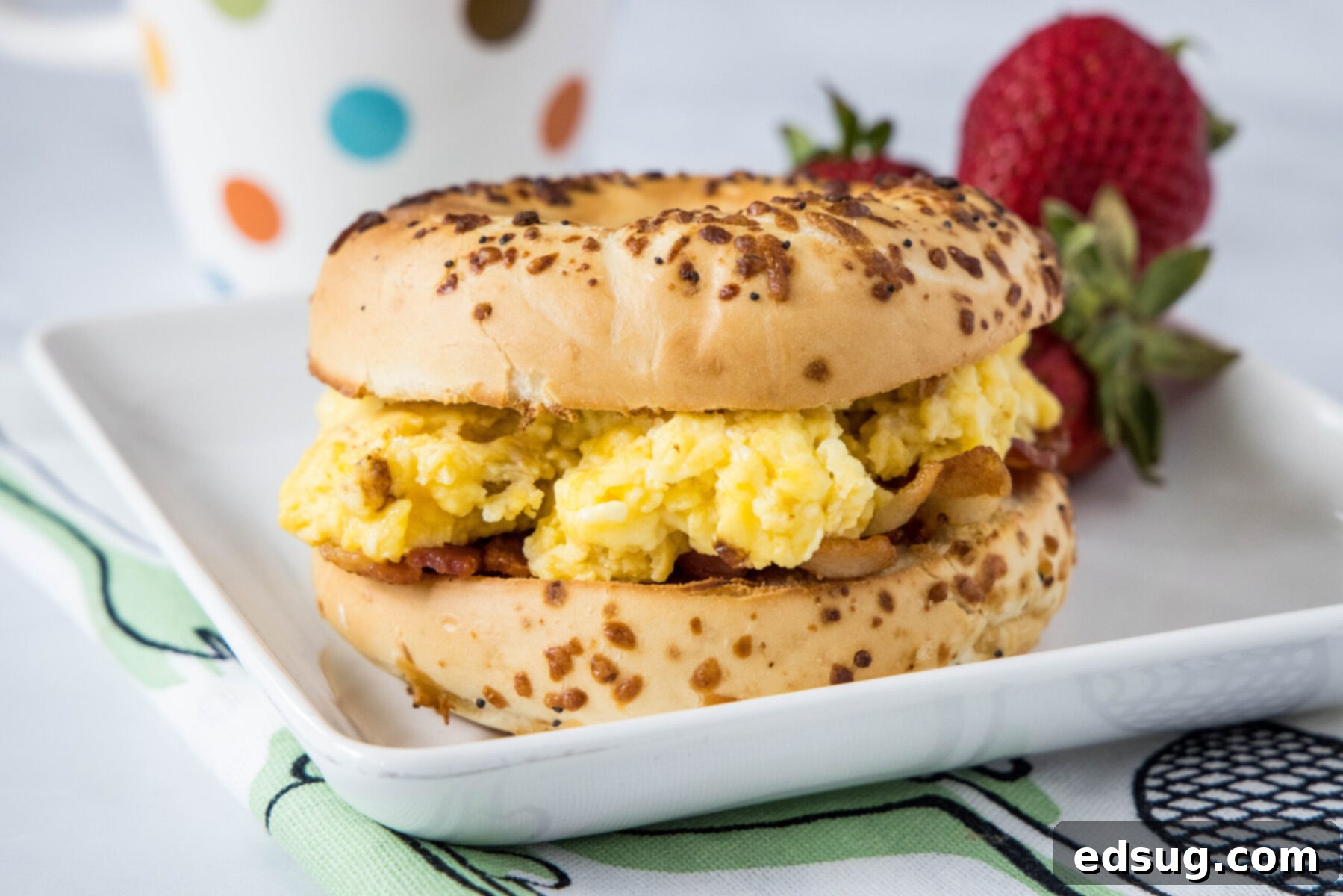 This classic bacon egg and cheese sandwich is the ultimate breakfast, with fluffy eggs, melty cheese, crispy bacon, and a toasted bagel. A bagel stuffed with eggs and bacon on a plate with strawberries and a mug in the background