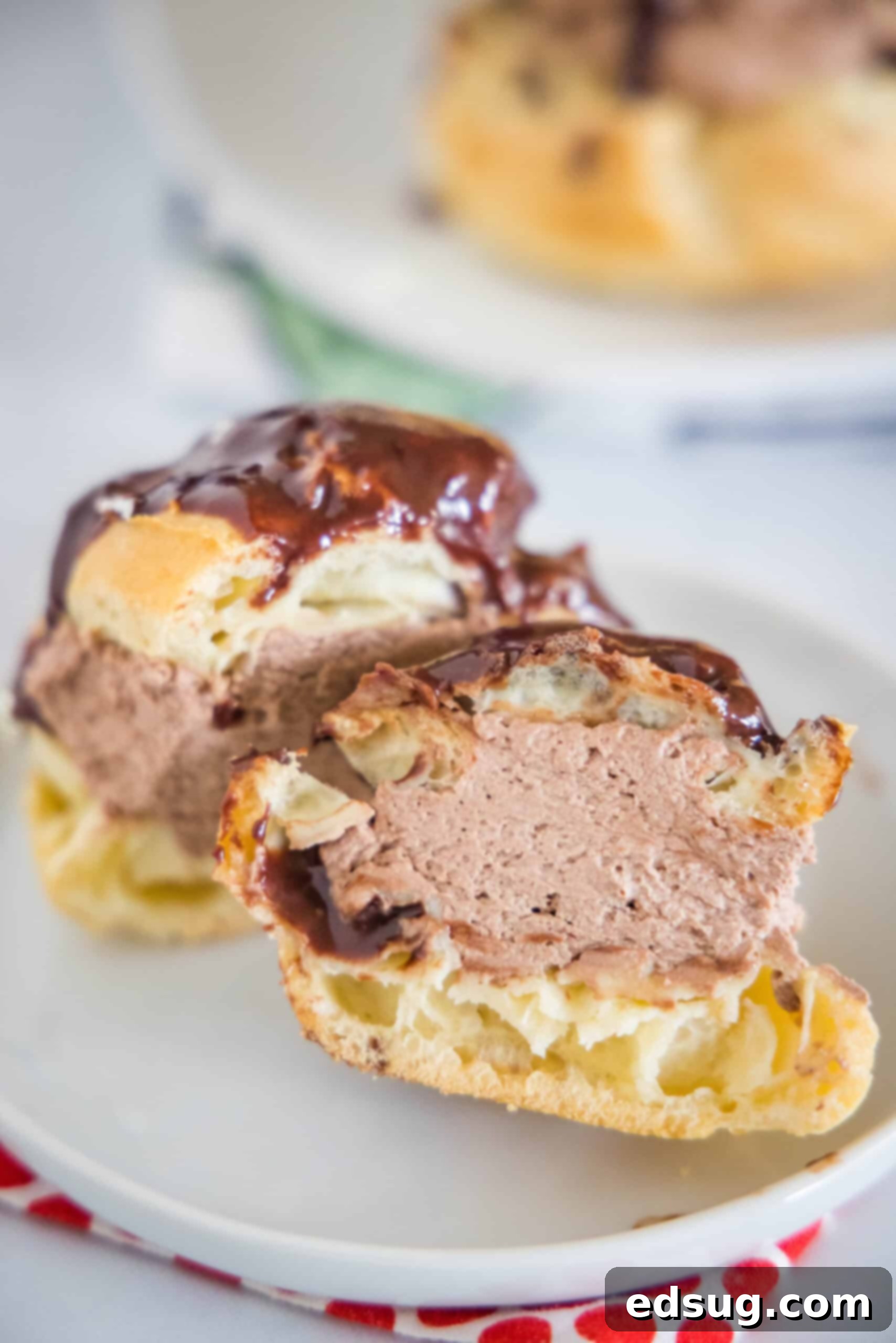 These chocolate cream puffs (profiteroles) are a chocolate lover’s dream, filled with fluffy chocolate whipped cream and topped with ganache. A perfectly cut chocolate cream puff, showcasing its fluffy chocolate whipped cream filling and glistening chocolate ganache topping, presented elegantly on a white plate.