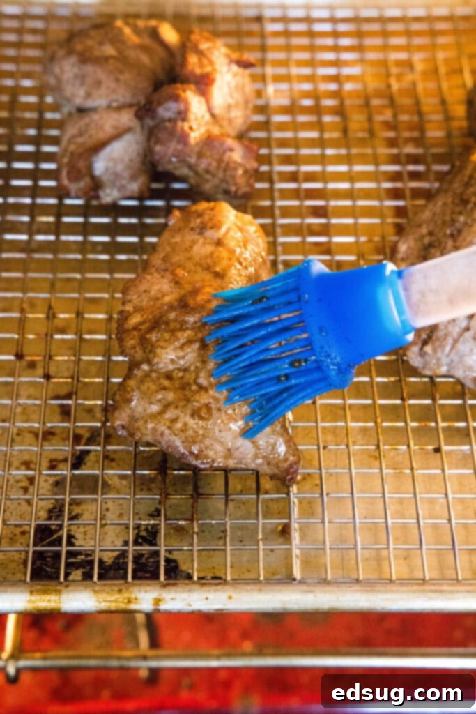 This easy char siu pork recipe is tender, juicy, and loaded with your favorite Chinese flavors like hoisin and five-spice powder.  A kitchen brush painting sauce on top of pieces of pork on a wire rack