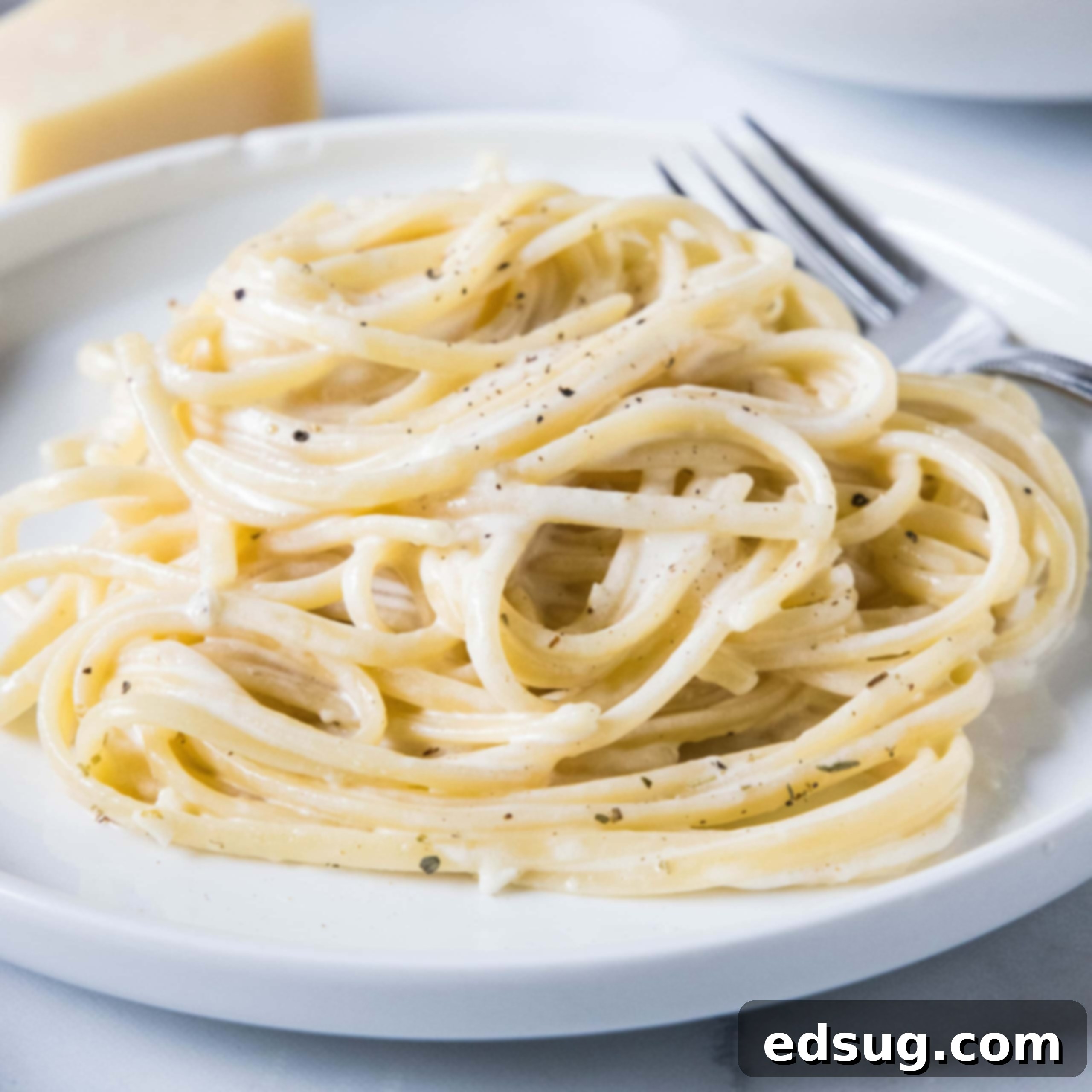 A serving of pasta alfredo on a white plate next to a fork, with a wedge of parmesan in the background.