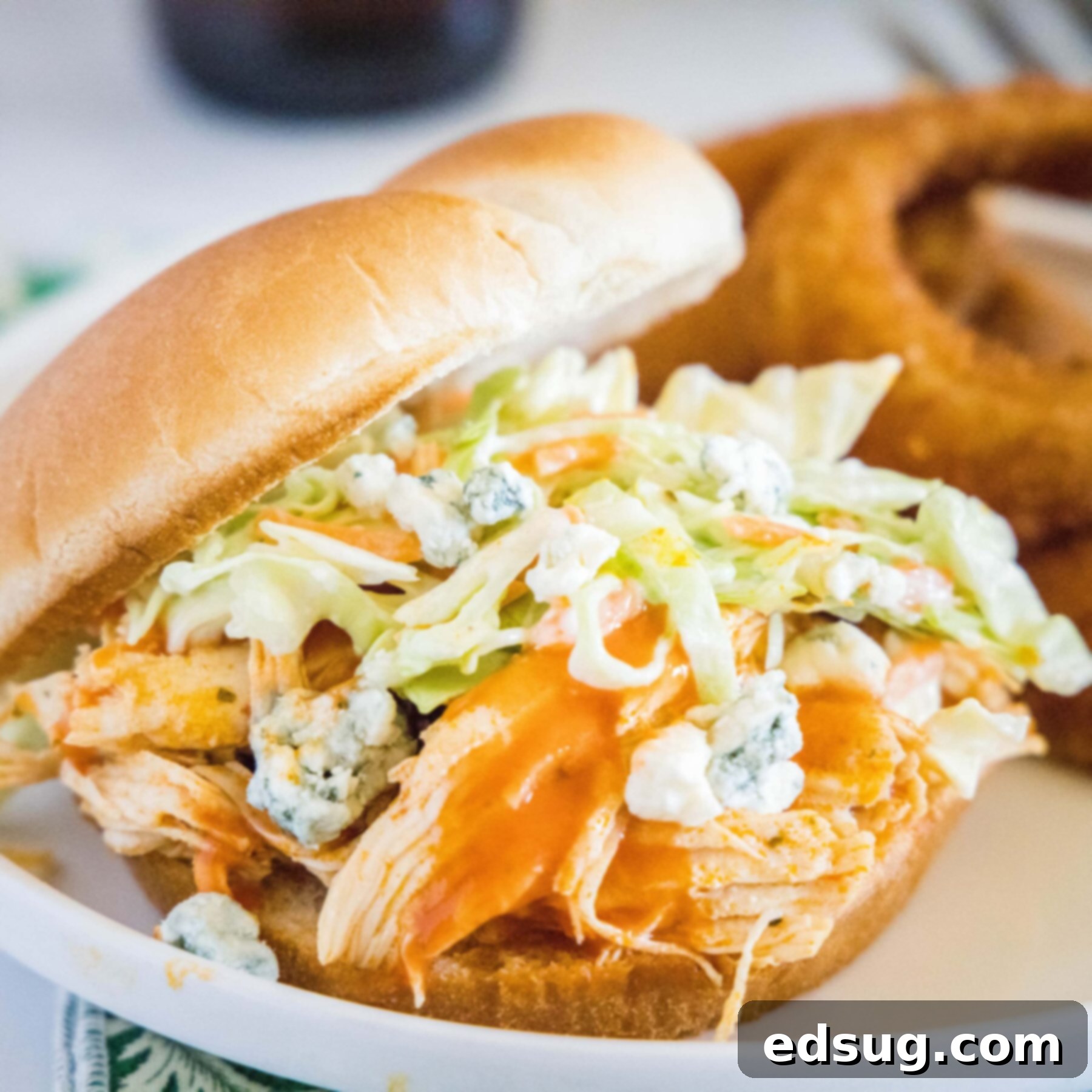 A buffalo chicken sandwich on a plate, topped with coleslaw and blue cheese, with onion rings in the background
