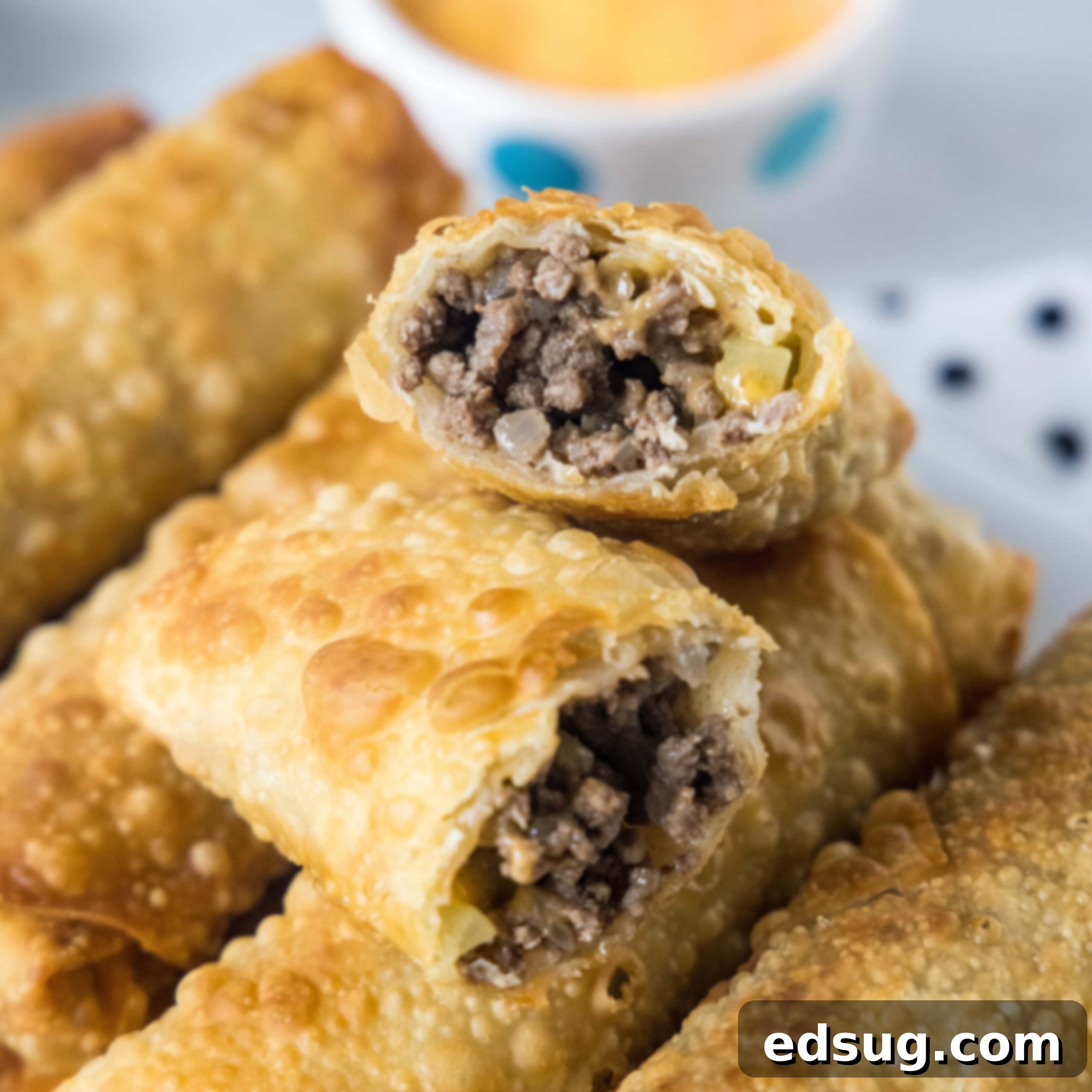 Close up of cheeseburger egg rolls stacked on a platter, with one egg roll broken in two and a bowl of dipping sauce in the background.
