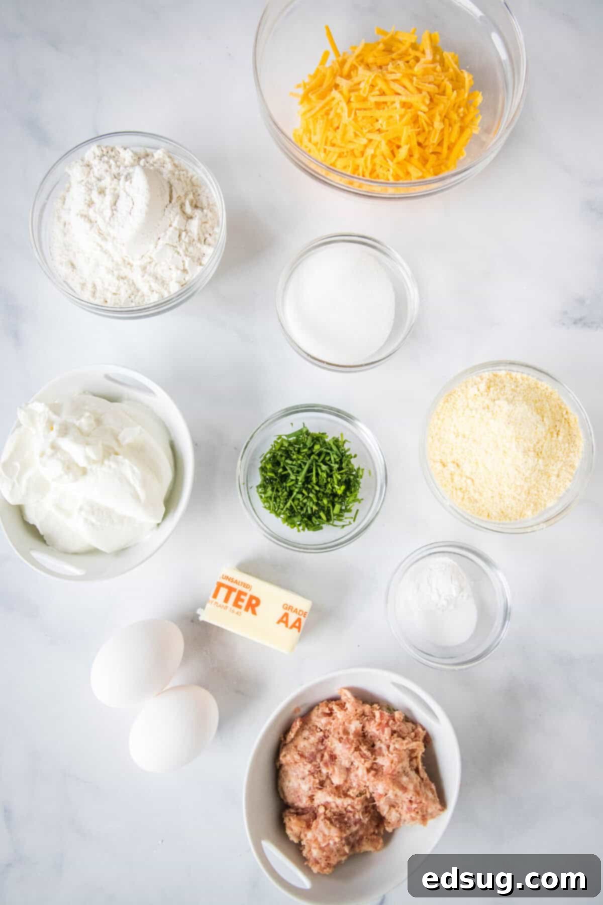 Cheesy Sausage Biscuit 2 Various ingredients for cheese and sausage biscuits displayed on a rustic wooden surface, including flour, sour cream, eggs, butter, browned sausage, shredded cheddar, and fresh chives.