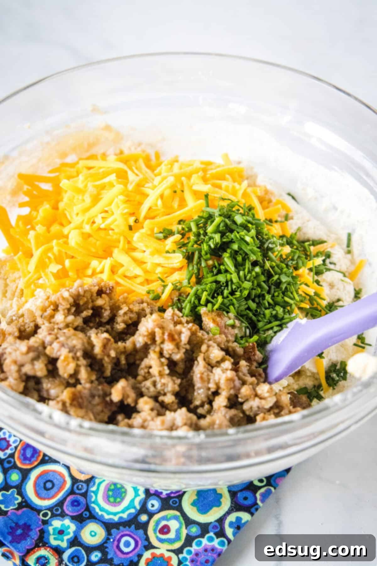 Cheesy Sausage Biscuit 3 Hands gently folding shredded cheddar cheese, browned crumbled sausage, and minced chives into a bowl of biscuit batter, ensuring even distribution.