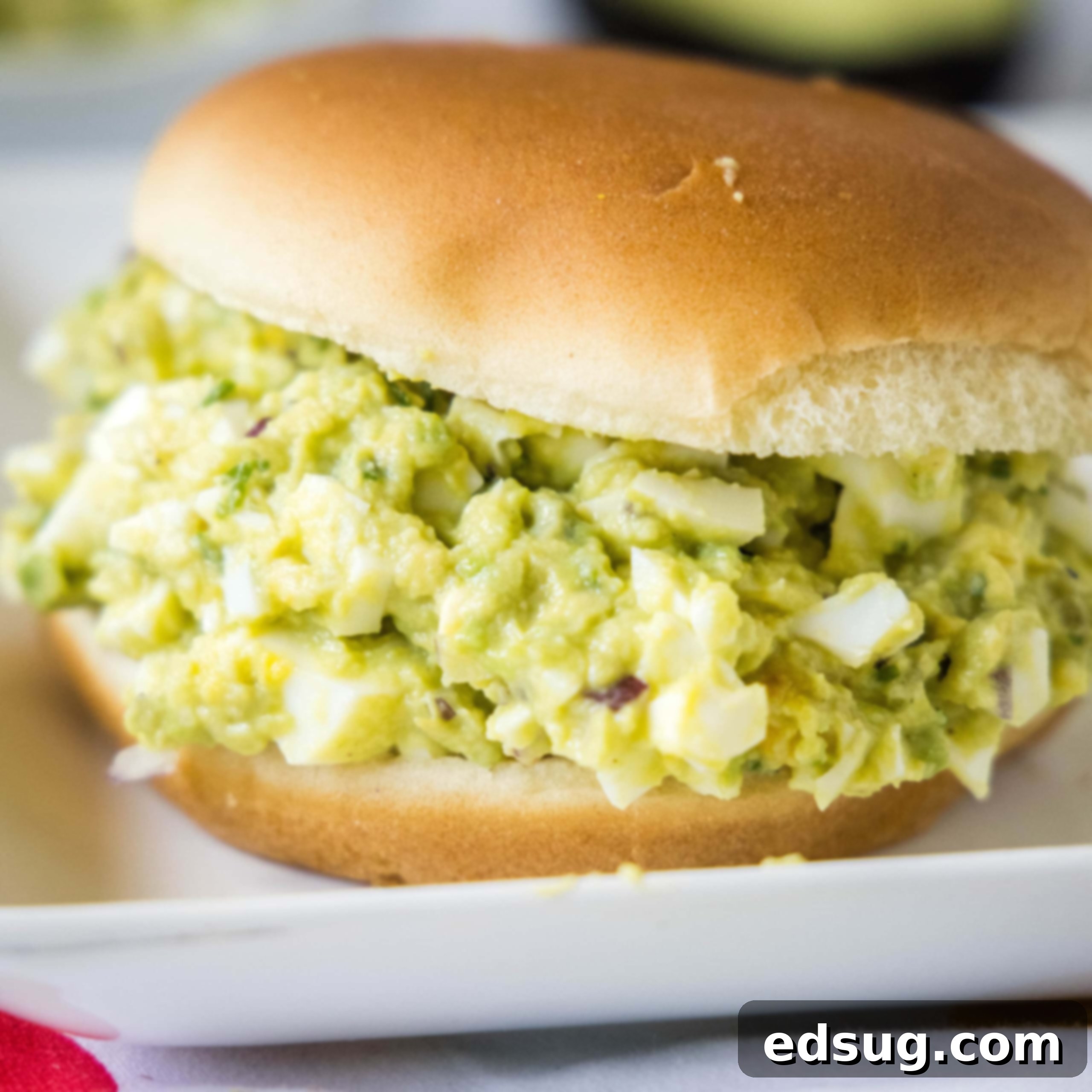Close up of an avocado egg salad sandwich on a toasted bun, garnished with fresh herbs.