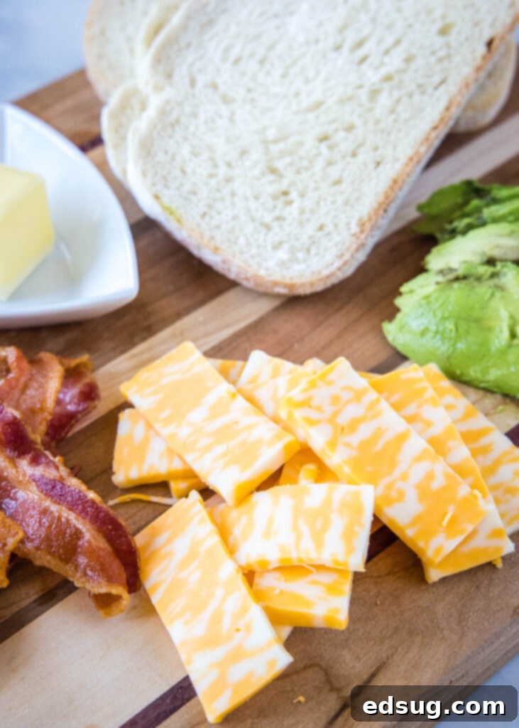 Ingredients for avocado grilled cheese sandwich, including bread, cheese, avocado, butter, and bacon, arranged on a cutting board.
