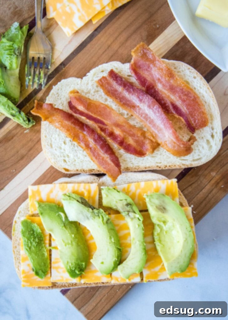 Assembling an avocado grilled cheese sandwich with cheese, avocado slices, and bacon on bread slices.