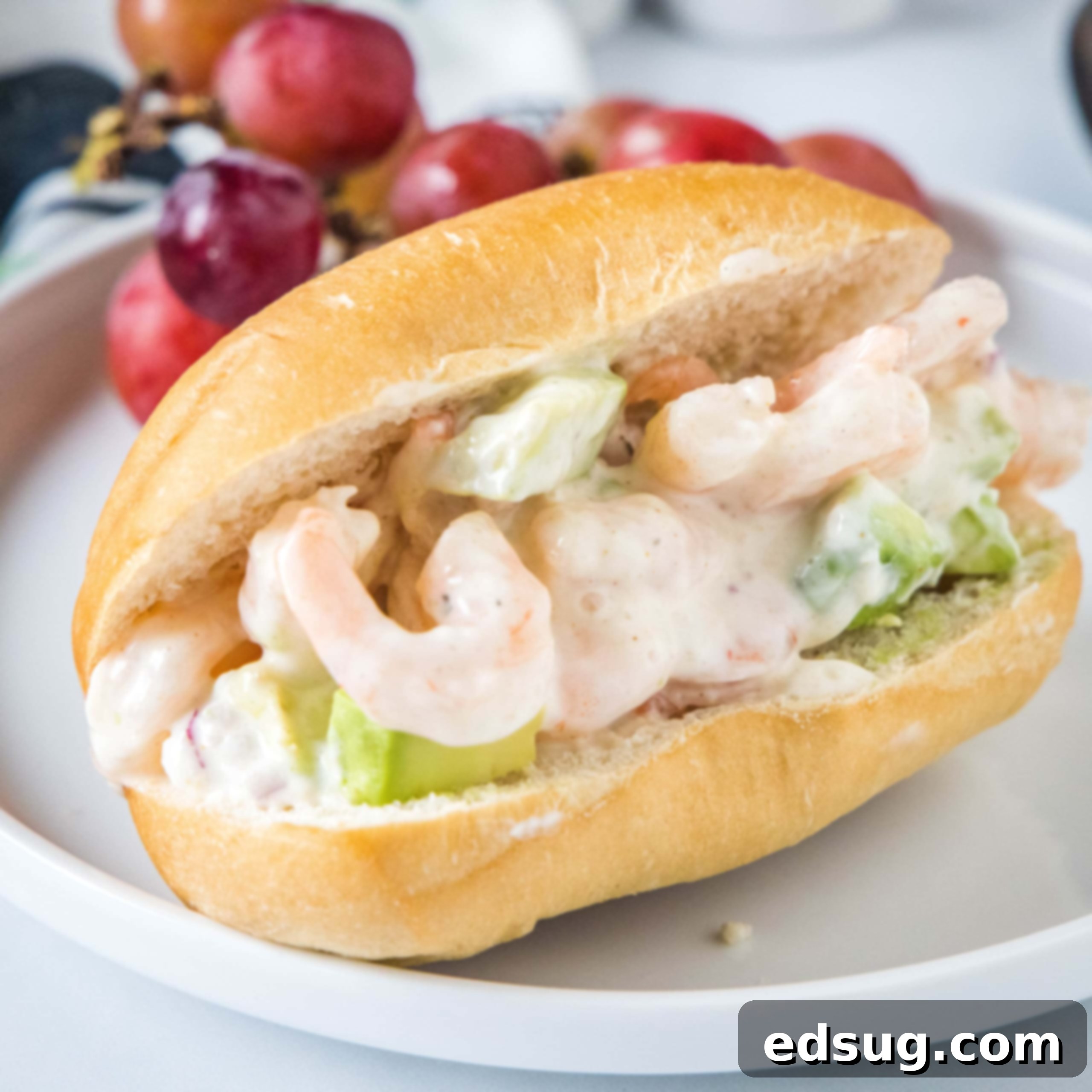 A fresh shrimp salad sandwich on a white plate with red grapes, perfect for lunch or a light dinner.