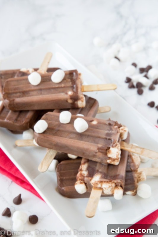 A trio of perfectly frozen hot chocolate popsicles, resting on a white plate, adorned with extra mini marshmallows.