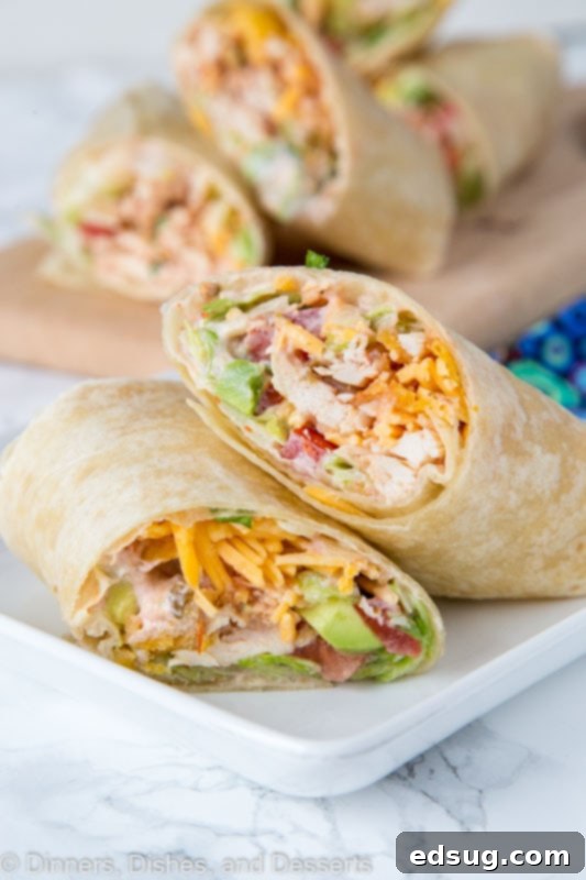 A chicken wrap cut in half, diagonally, with one half leaning against the other, on a plate, with a cutting board full of wraps in the background