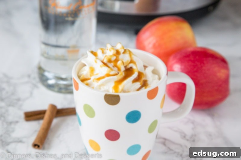 Boozy Warm Apple Cider 5 A wide shot of a glass mug filled with spiked hot apple cider, topped with whipped cream and caramel, next to fresh apples and cinnamon sticks.