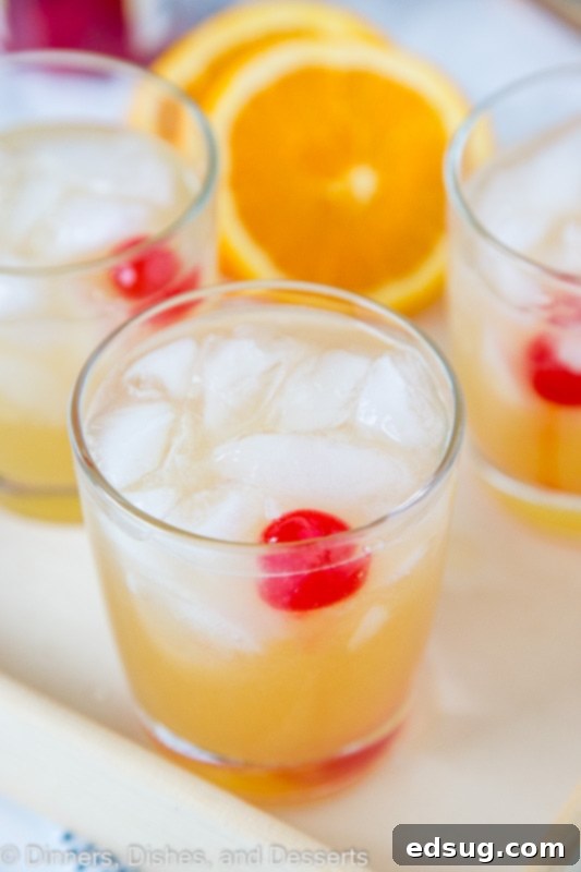 Two glasses of Whiskey Sour, one garnished with a maraschino cherry and orange slice, the other with a cherry, both filled with ice, on a stylish tray.