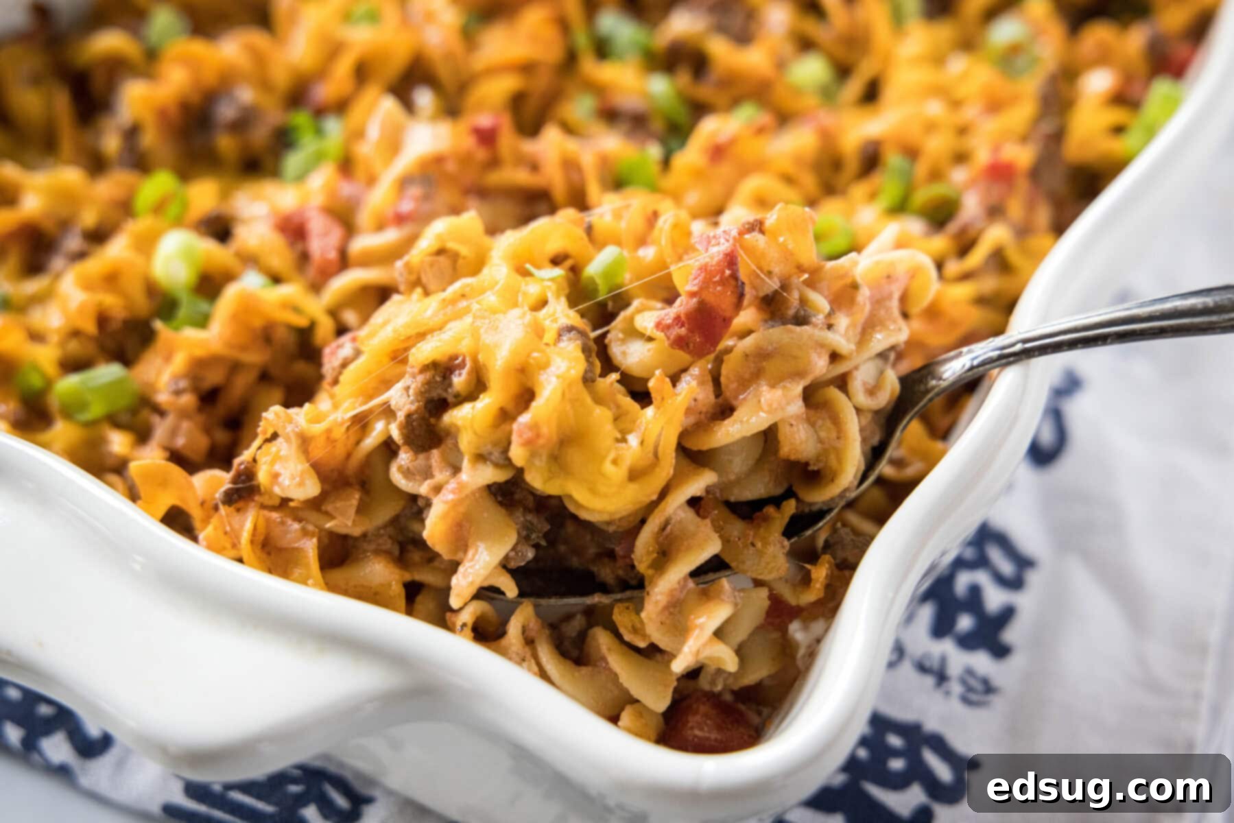 Easy beef noodle casserole is packed with juicy meat, a hearty and creamy sauce, tender egg noodles, and melted cheddar cheese. The corner of a casserole dish filled with beef noodle casserole, with a serving spoon