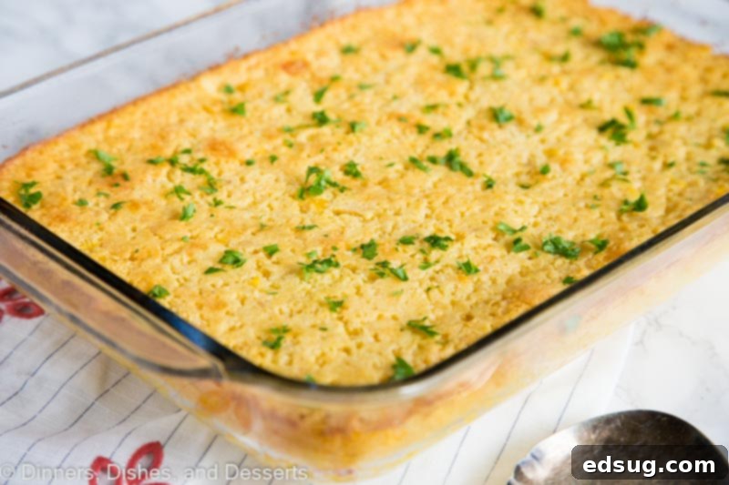 A full baking dish of freshly baked corn casserole, golden brown and bubbling slightly, with a spoon ready for serving.
