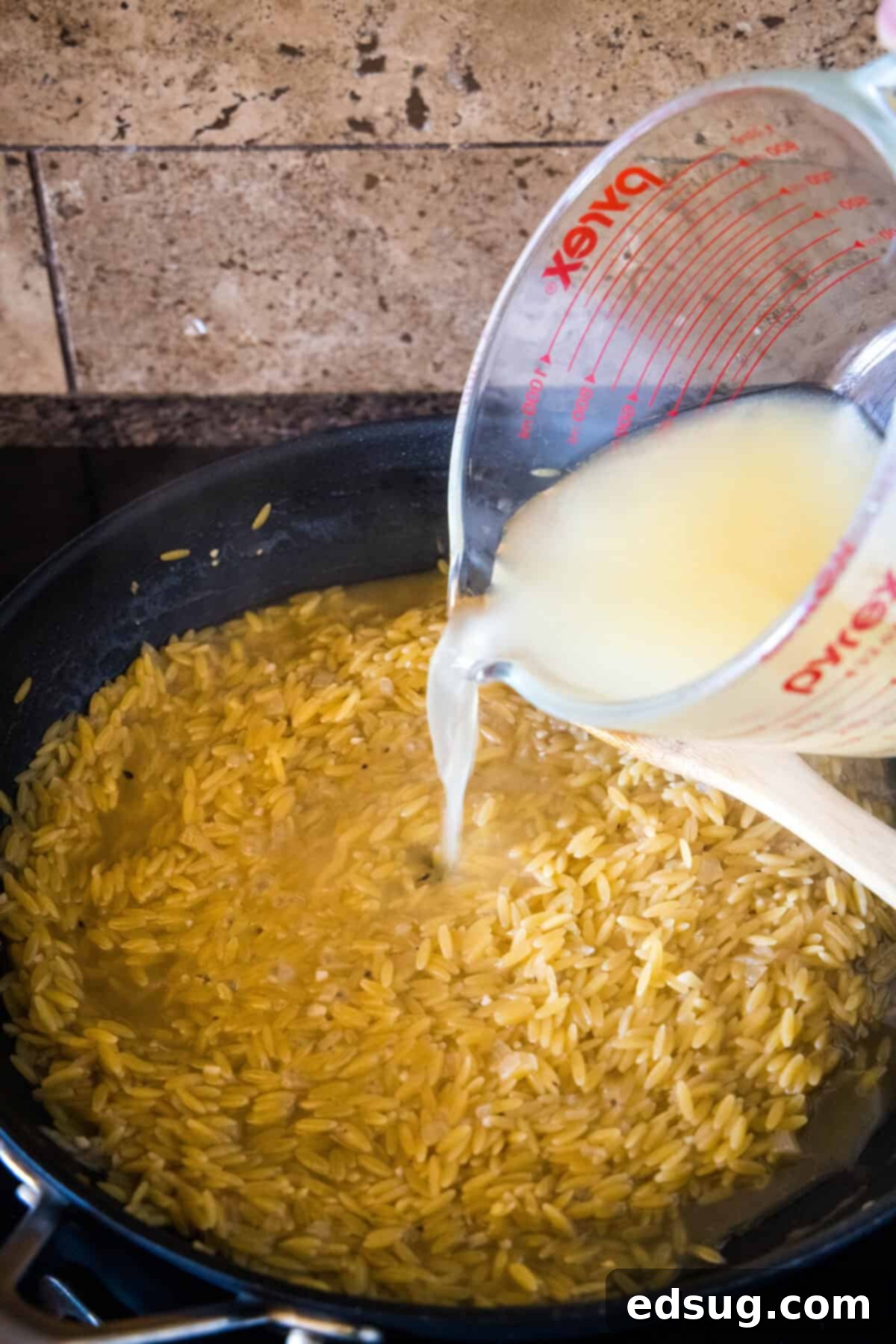 Cooking the orzo pasta by gradually adding chicken broth in the same skillet. Chicken broth being slowly poured into a skillet with sautéed dried orzo and onions.