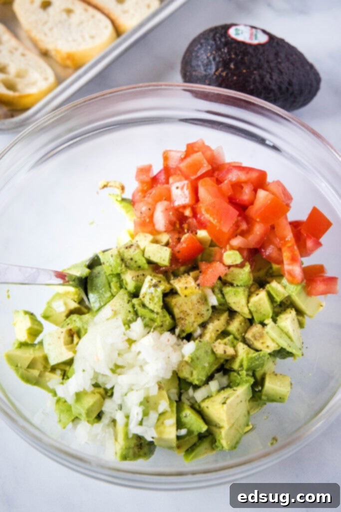 Close-up of a vibrant avocado, tomato, and red onion mixture in a white mixing bowl, ready to top the bruschetta.