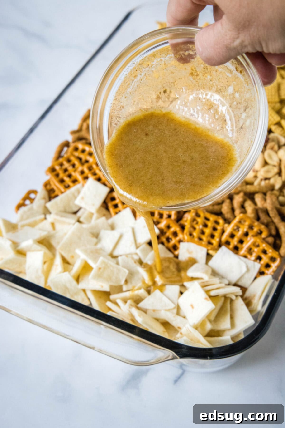 Mexican Snack Mix 3 A close-up of a baking dish where melted butter and spice mixture are being poured over a medley of snack ingredients, illustrating the coating process.