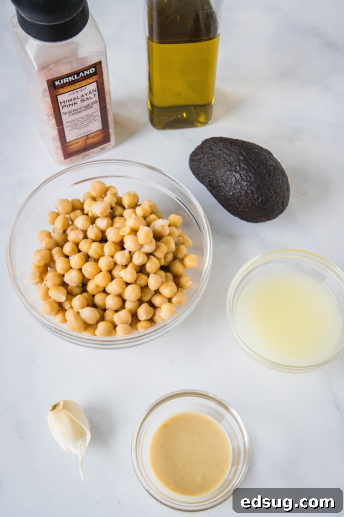 All the fresh ingredients for avocado hummus laid out on a cutting board: avocado, chickpeas, lemon, garlic, olive oil, and tahini.
