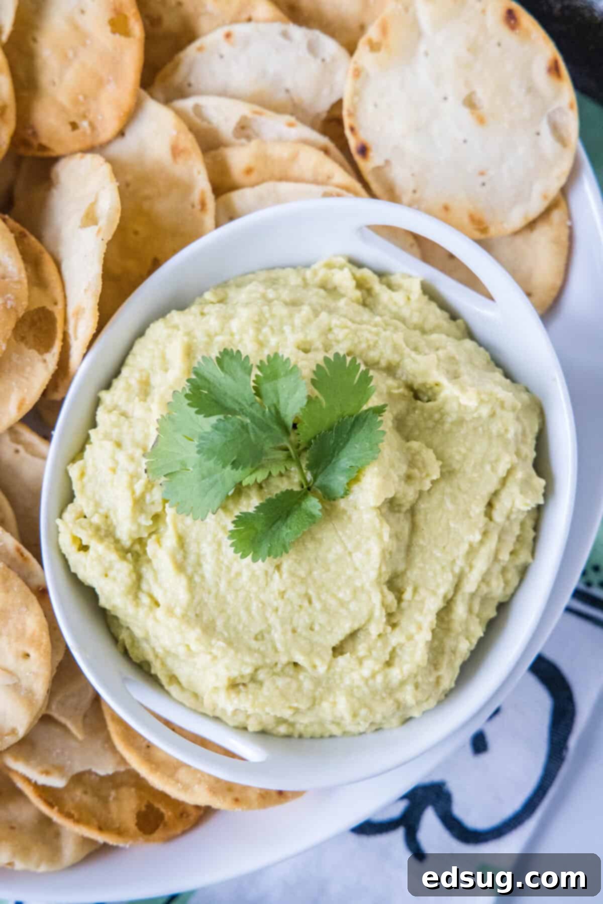 Overhead shot of a bowl of avocado hummus garnished with a swirl of olive oil and a sprinkle of fresh herbs.