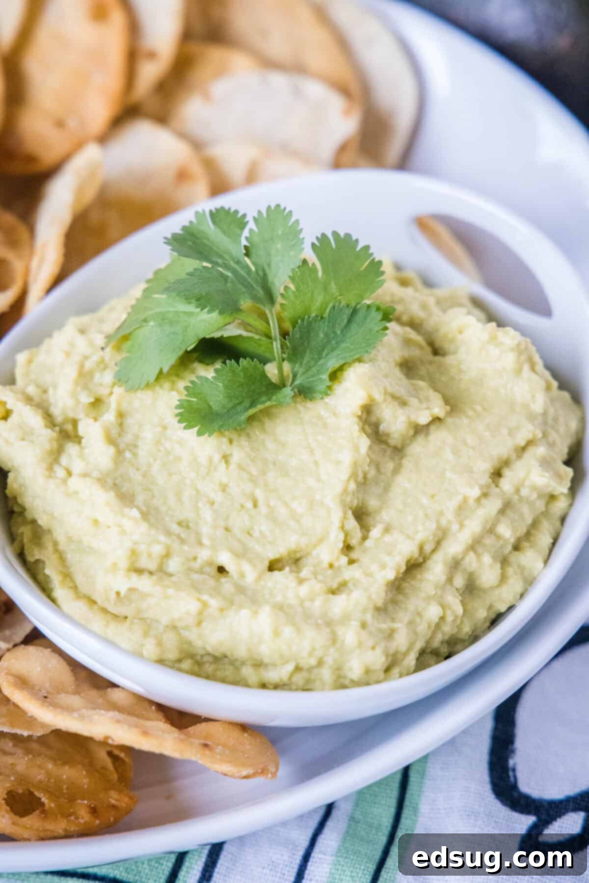 A white bowl with smooth avocado hummus, topped with a vibrant sprig of cilantro and a drizzle of olive oil.