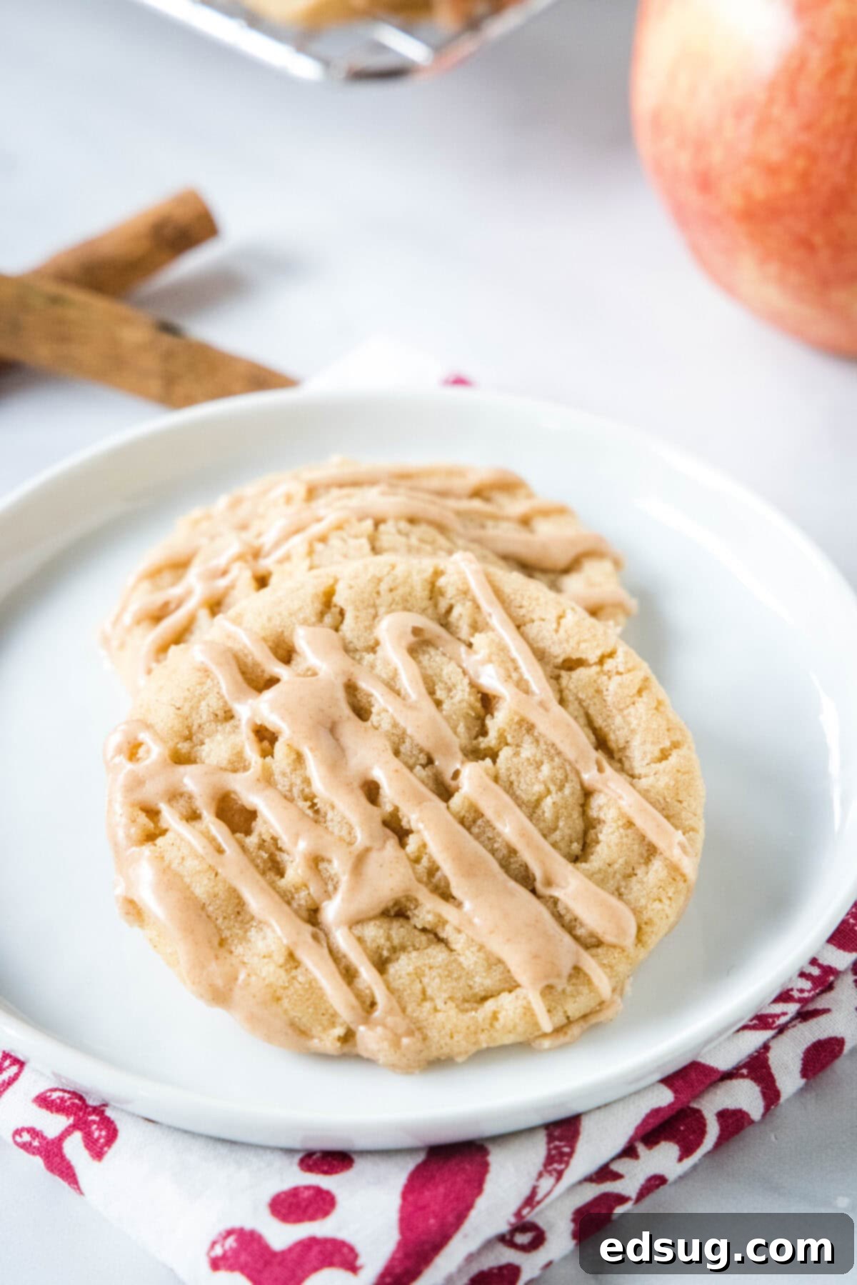 Apple Cider Cookies 2_labled Two apple cider cookies drizzled with cider glaze on a white plate, with an apple and cinnamon sticks in the background.
