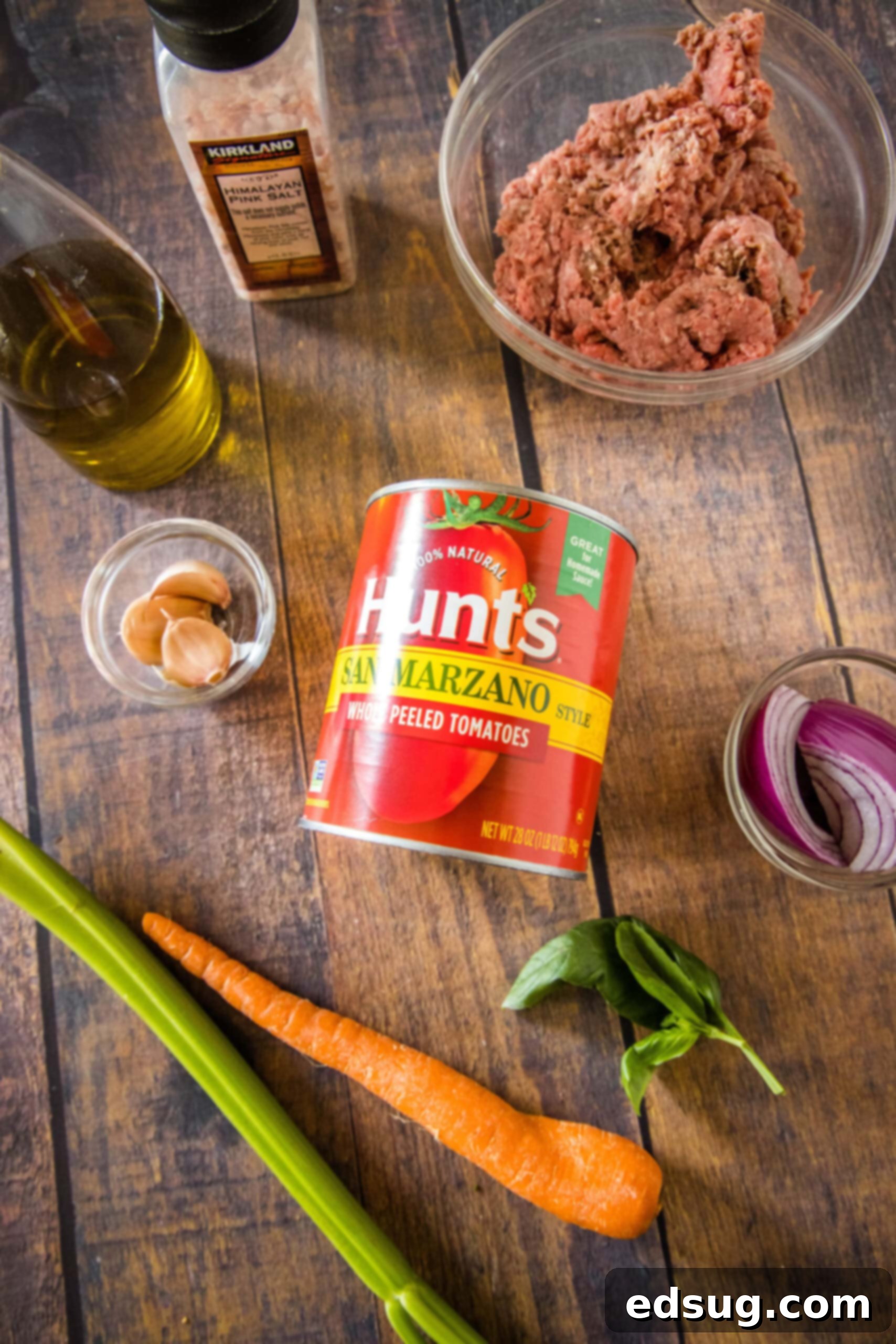 A rustic wooden cutting board displaying the fresh, high-quality ingredients required for preparing a delicious beef ragu: carrots, celery, onion, garlic, ground beef, and San Marzano tomato sauce.