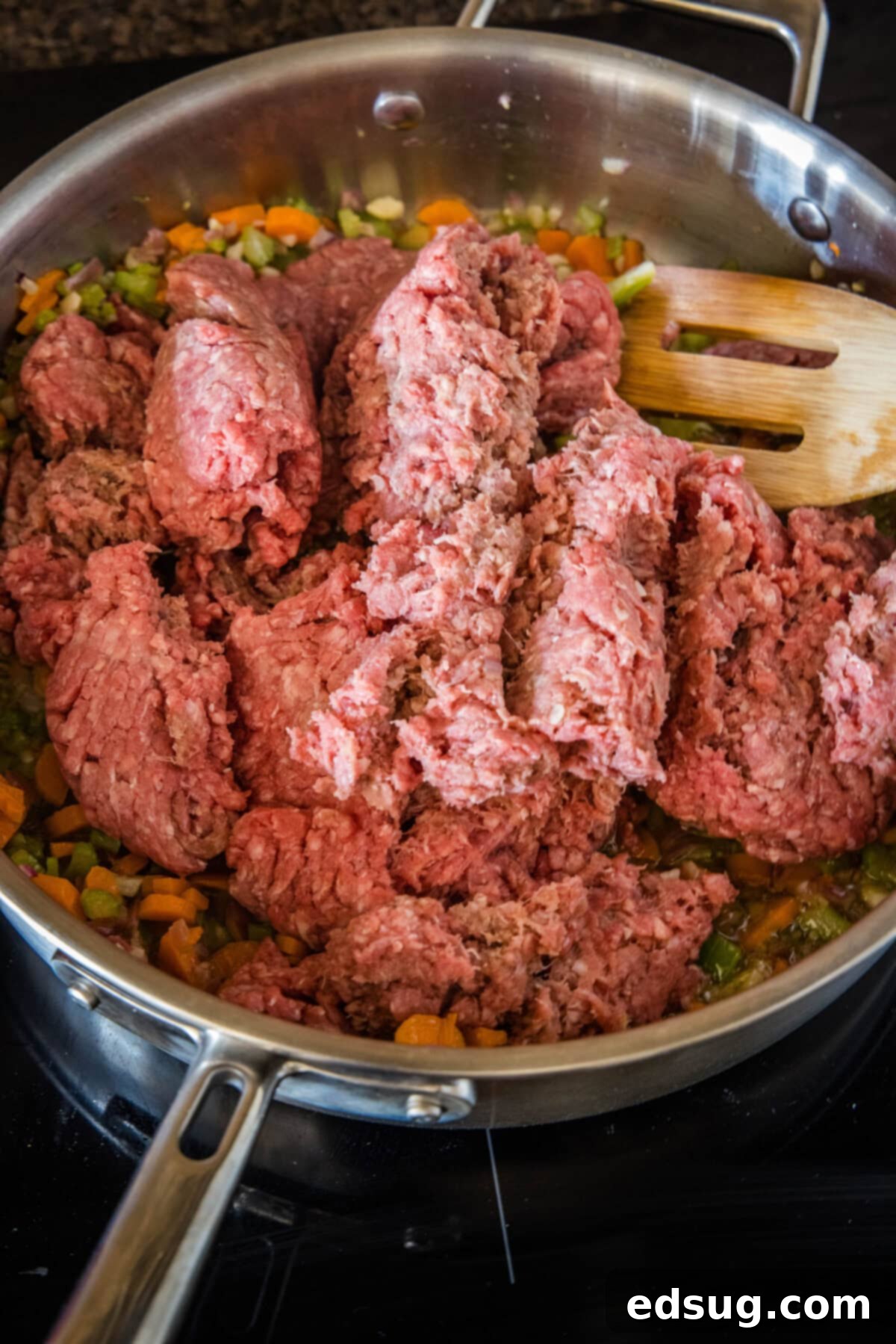Lean ground beef beginning to brown and break apart in a skillet with a wooden spatula, blending with the sautéed vegetables.