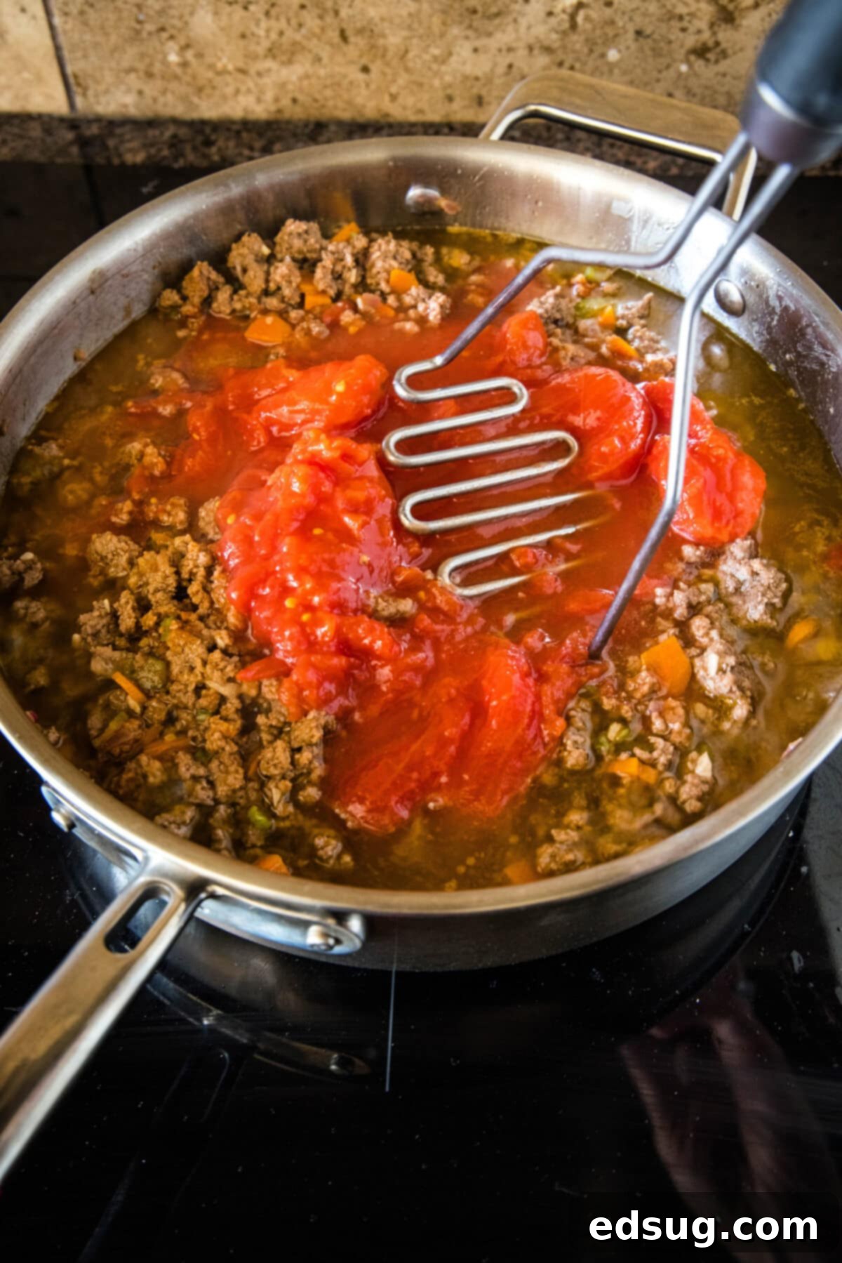 Crushed San Marzano tomatoes being stirred into the browned ground beef and sautéed vegetables in a large skillet, forming the base of the rich ragu sauce.