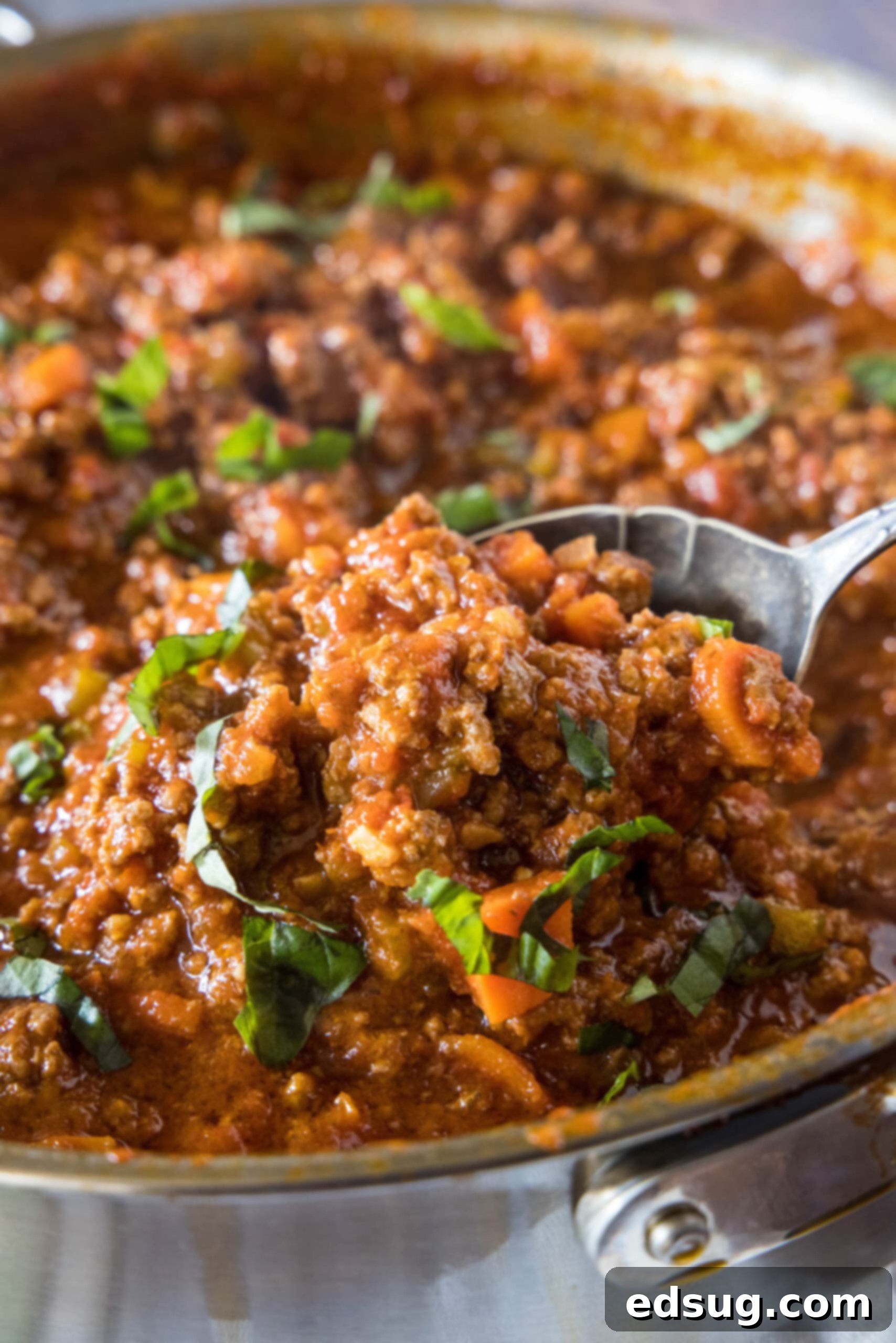 A wooden spoon lifting a spoonful of rich, chunky beef ragu from a large, rustic deep skillet, showcasing its hearty texture.