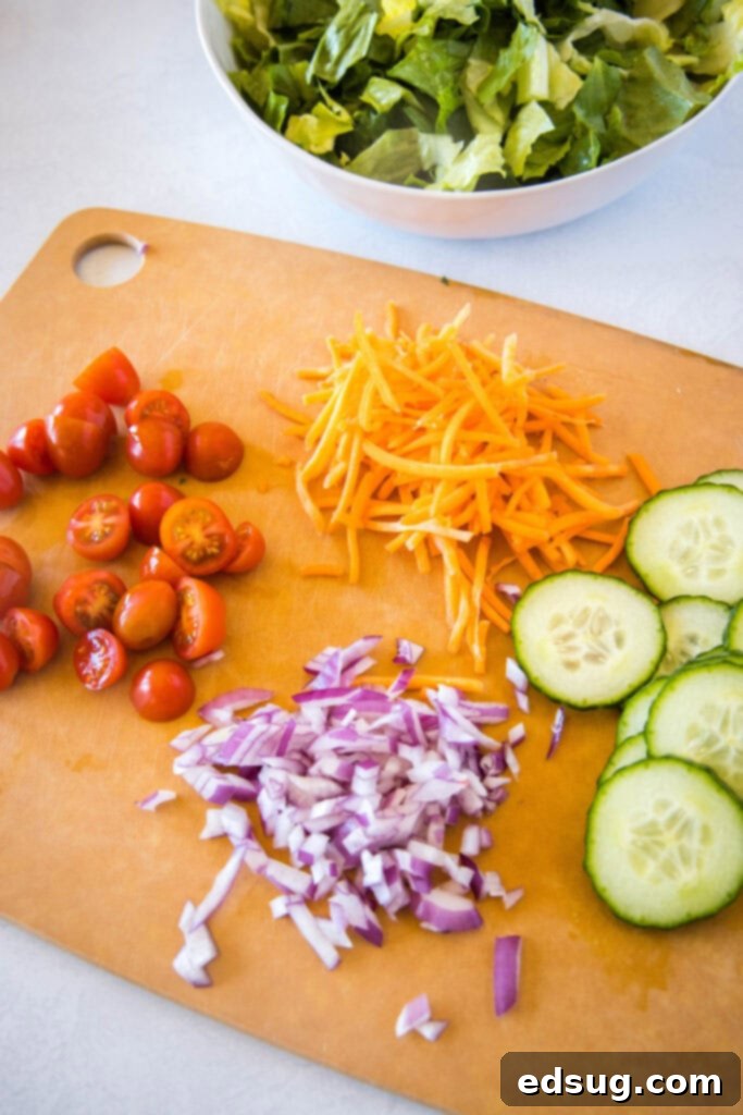 A selection of fresh side salad ingredients including lettuce, cucumber, grape tomatoes, and red onion, laid out on a cutting board.