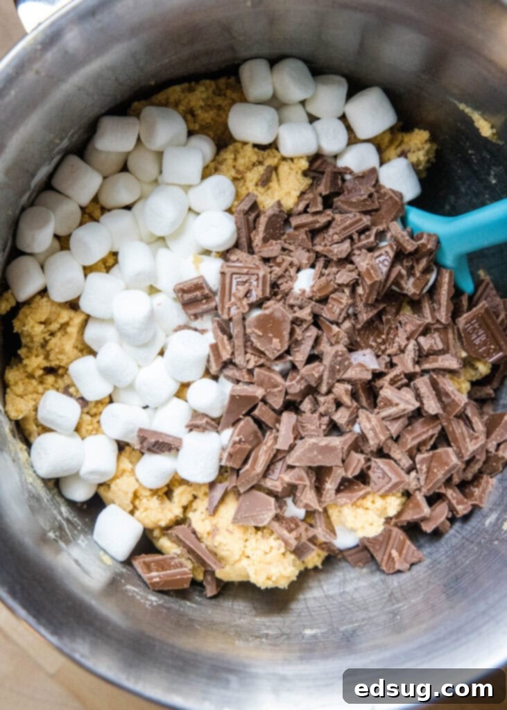 Smores Cookies 5 Close-up of cookie dough in a mixing bowl, generously topped with chocolate chips and mini marshmallows, ready to be mixed.
