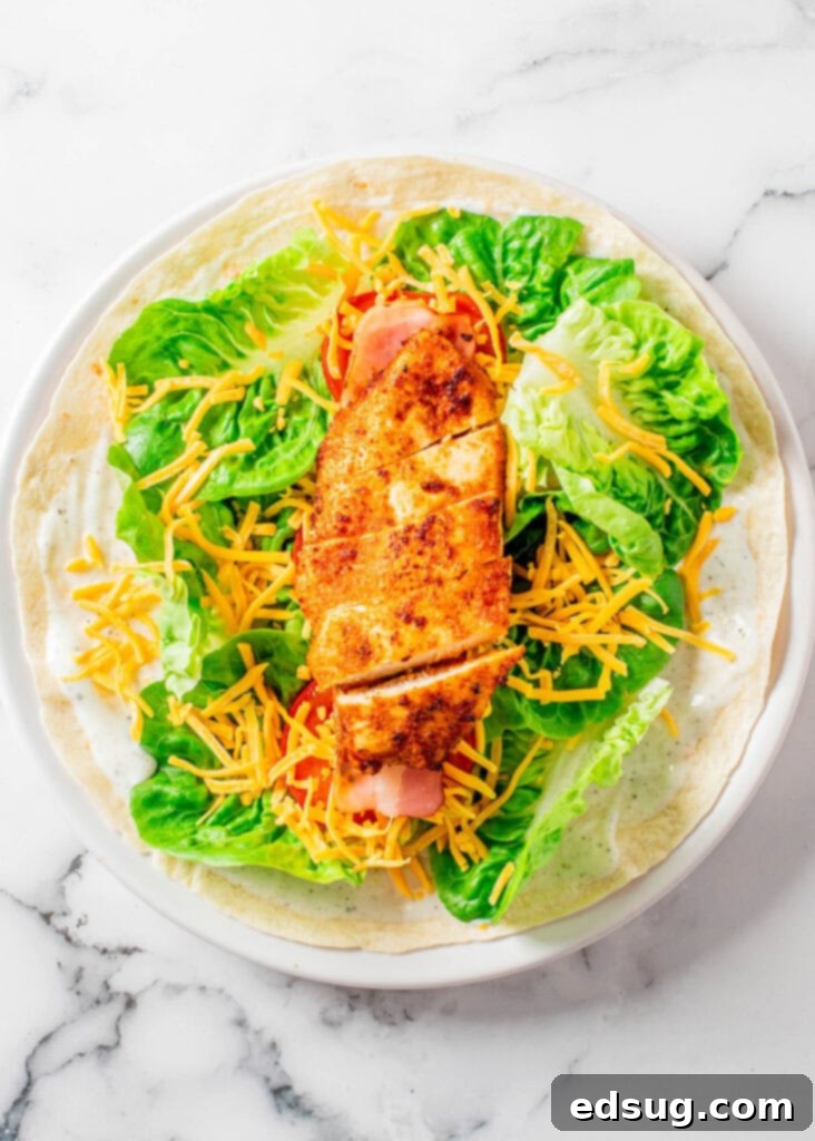Signature Chicken Bacon Ranch Wrap 3 Close-up of hands assembling a chicken bacon ranch wrap on a white plate with ingredients visible.