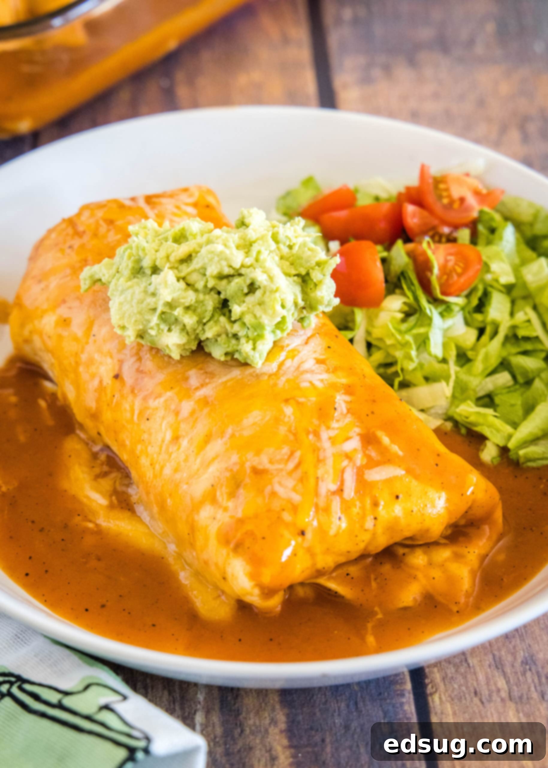 A mouthwatering wet beef burrito topped with guacamole, served with a side salad. A wet burrito on a white plate, topped with guacamole, next to fresh lettuce and tomatoes