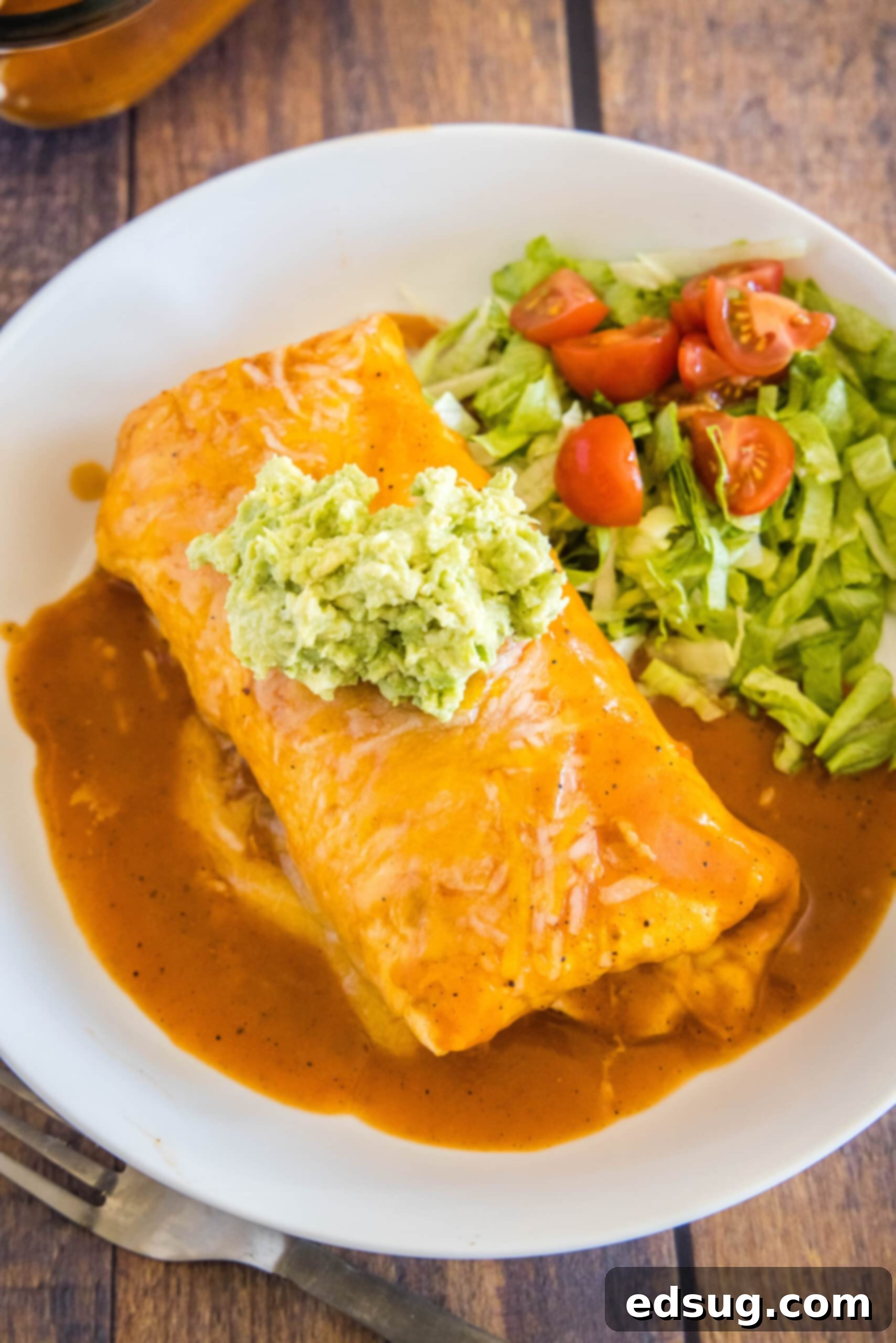 An overhead shot of a saucy wet burrito, garnished with fresh guacamole, alongside a fork and a crisp salad. Overhead view of a wet burrito topped with guacamole, on a plate with lettuce and tomatoes, next to a fork