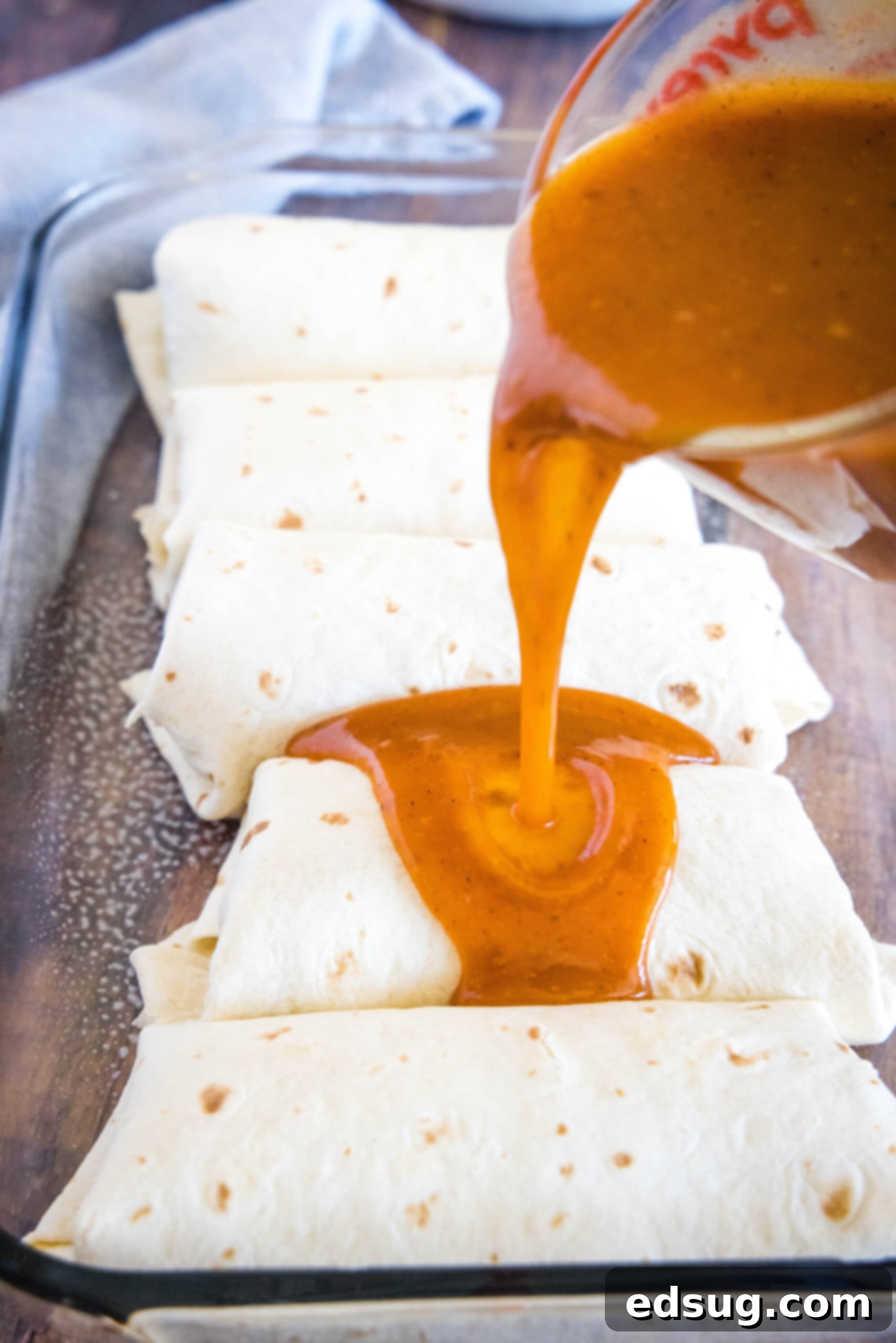 Pouring savory enchilada sauce over freshly assembled burritos in a baking dish. Enchilada sauce being poured over burritos nestled in a baking dish.