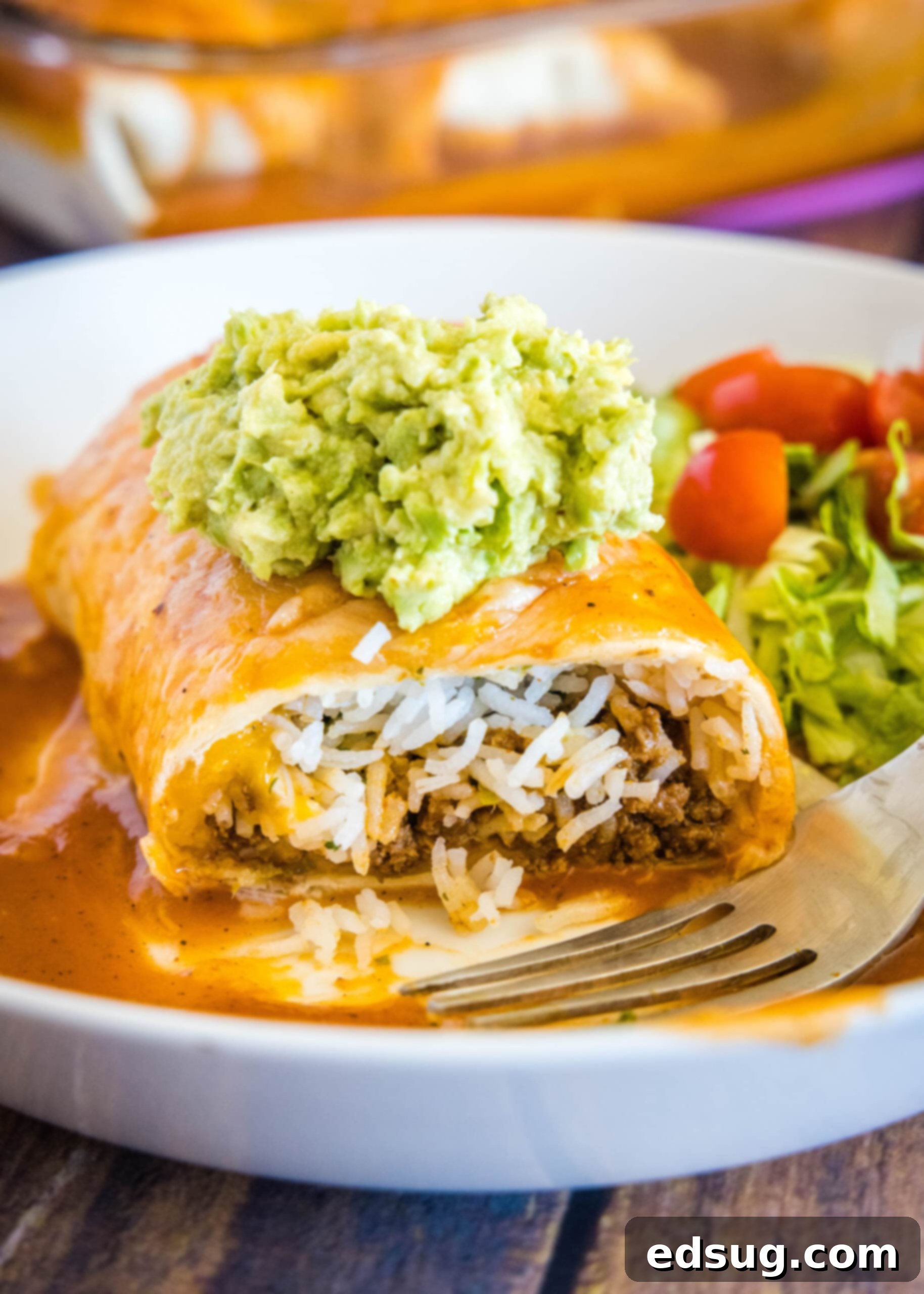 A cross-section of a wet beef burrito, revealing its flavorful filling, served with guacamole and a fresh salad. Cross section of half a wet burrito topped with guacamole on a plate, with beef and rice spilling out of it, next to a fork, some lettuce, and some tomato slices.