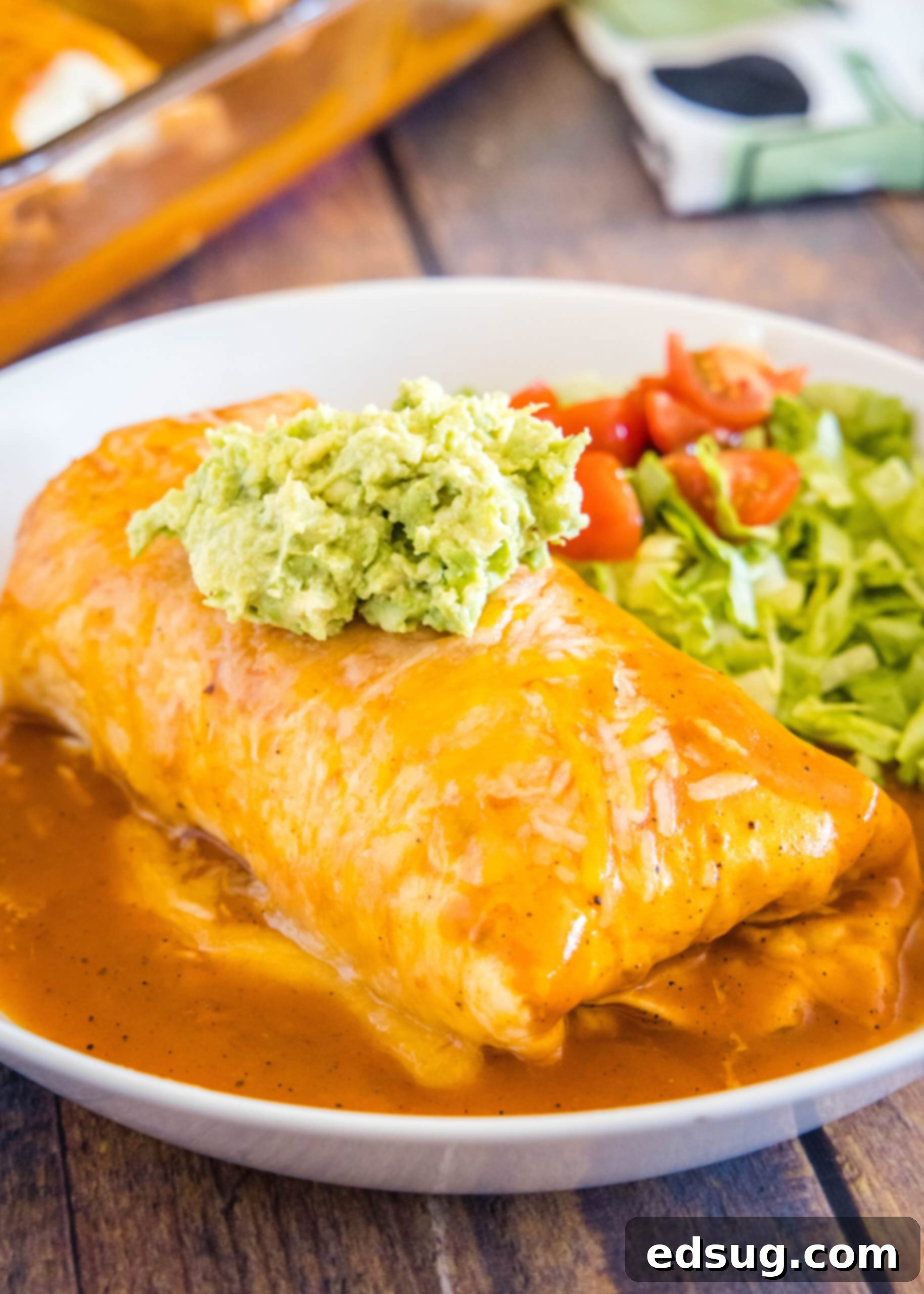 A perfectly prepared wet burrito, generously topped with guacamole, served with a fresh salad. A wet burrito on a plate topped with guacamole, with a salad of lettuce and tomatoes in the background.