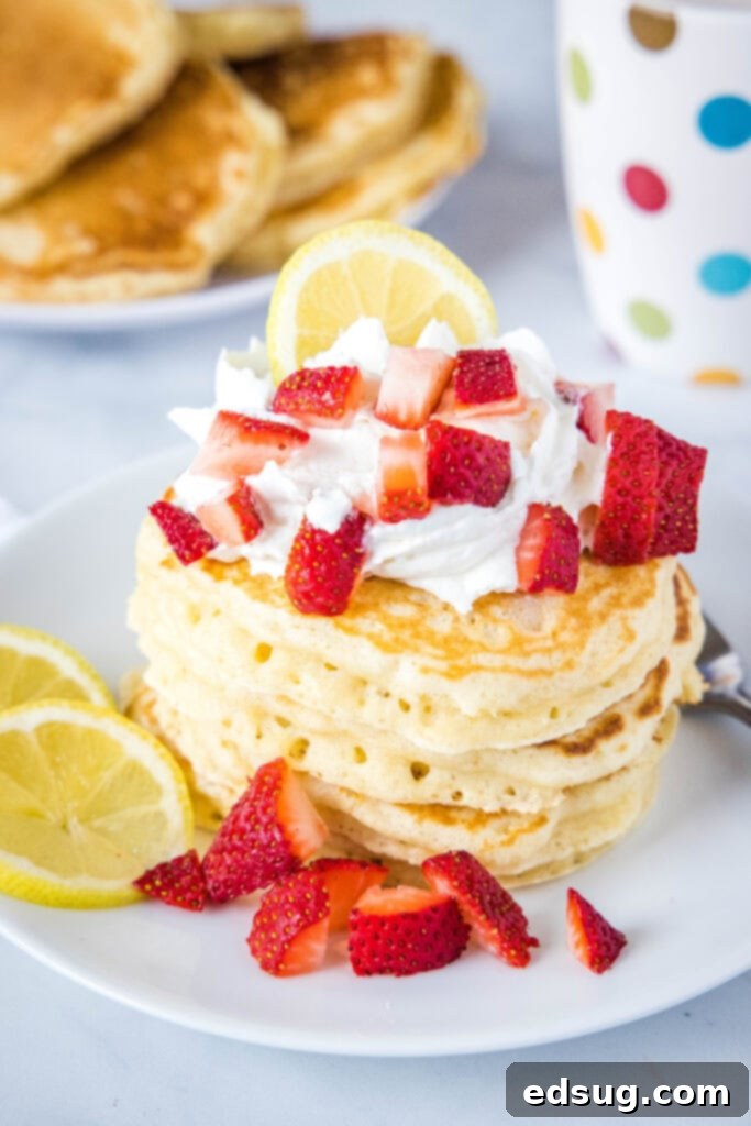 Tall stack of golden lemon pancakes on a white plate, garnished with whipped cream and sliced strawberries