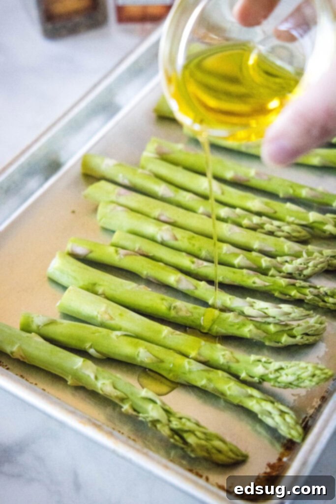 Olive oil being drizzled generously over fresh, trimmed asparagus spears spread out on a baking sheet, ready for seasoning.