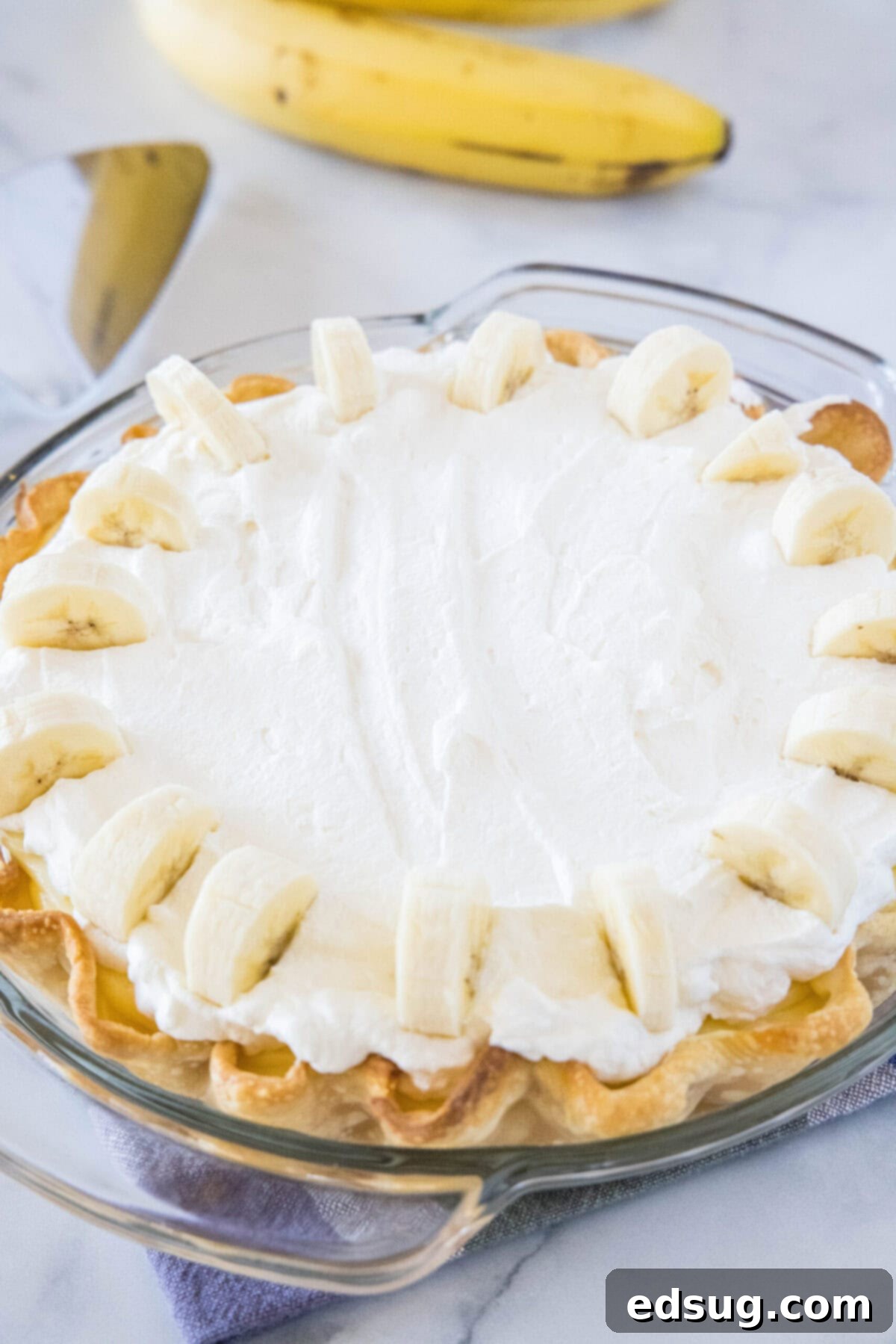 A perfectly set banana cream pie, garnished with fresh banana slices, with a whole banana in the soft-focus background, highlighting its key ingredient.