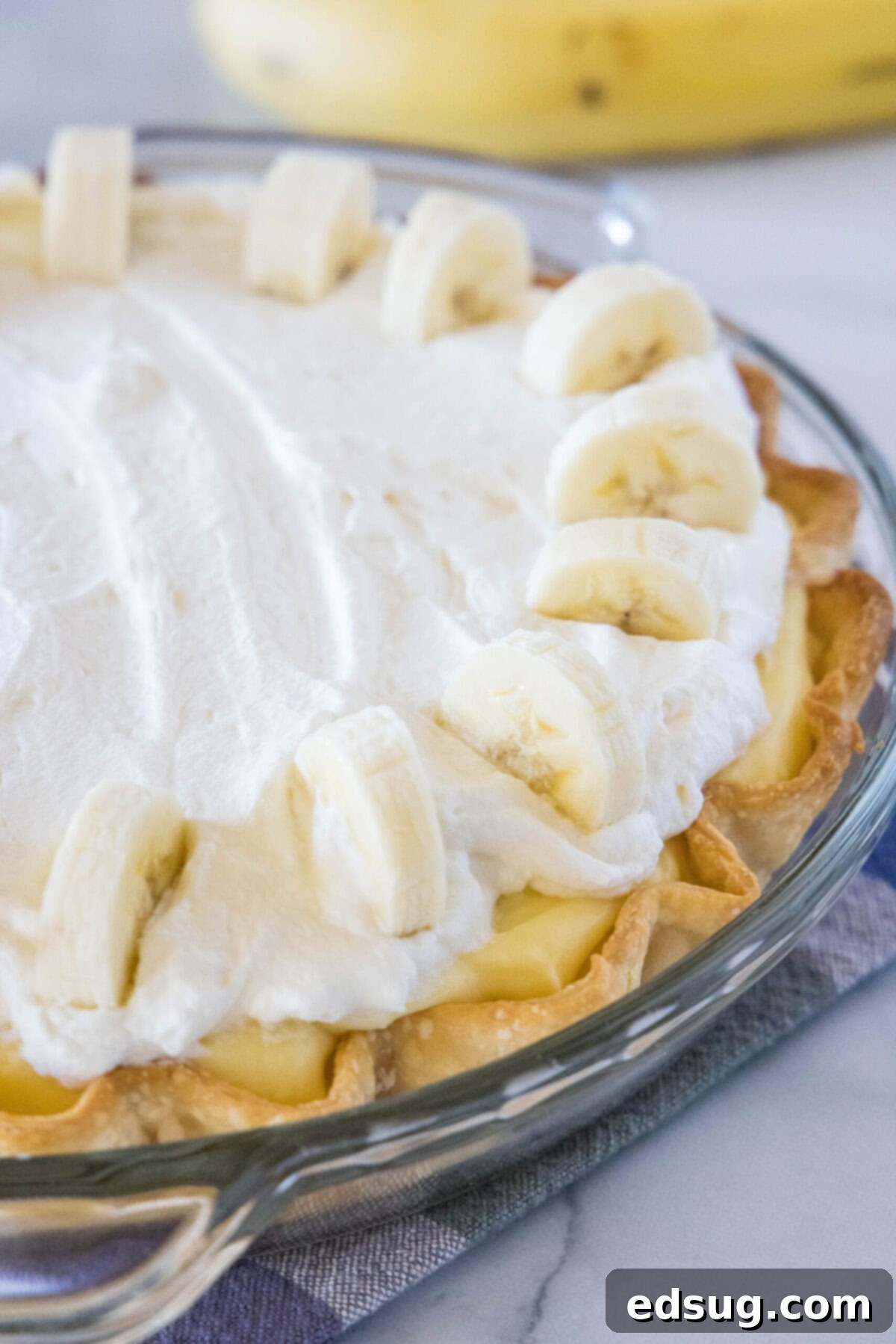 A beautiful close-up of a finished banana cream pie, revealing the layers of golden crust, creamy banana custard, and swirls of whipped cream, garnished with banana slices.