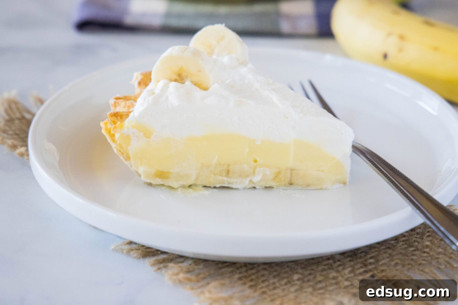 A delectable slice of banana cream pie on a pristine white plate, accompanied by a fork, with a blurred fresh banana in the background, ready to be enjoyed.