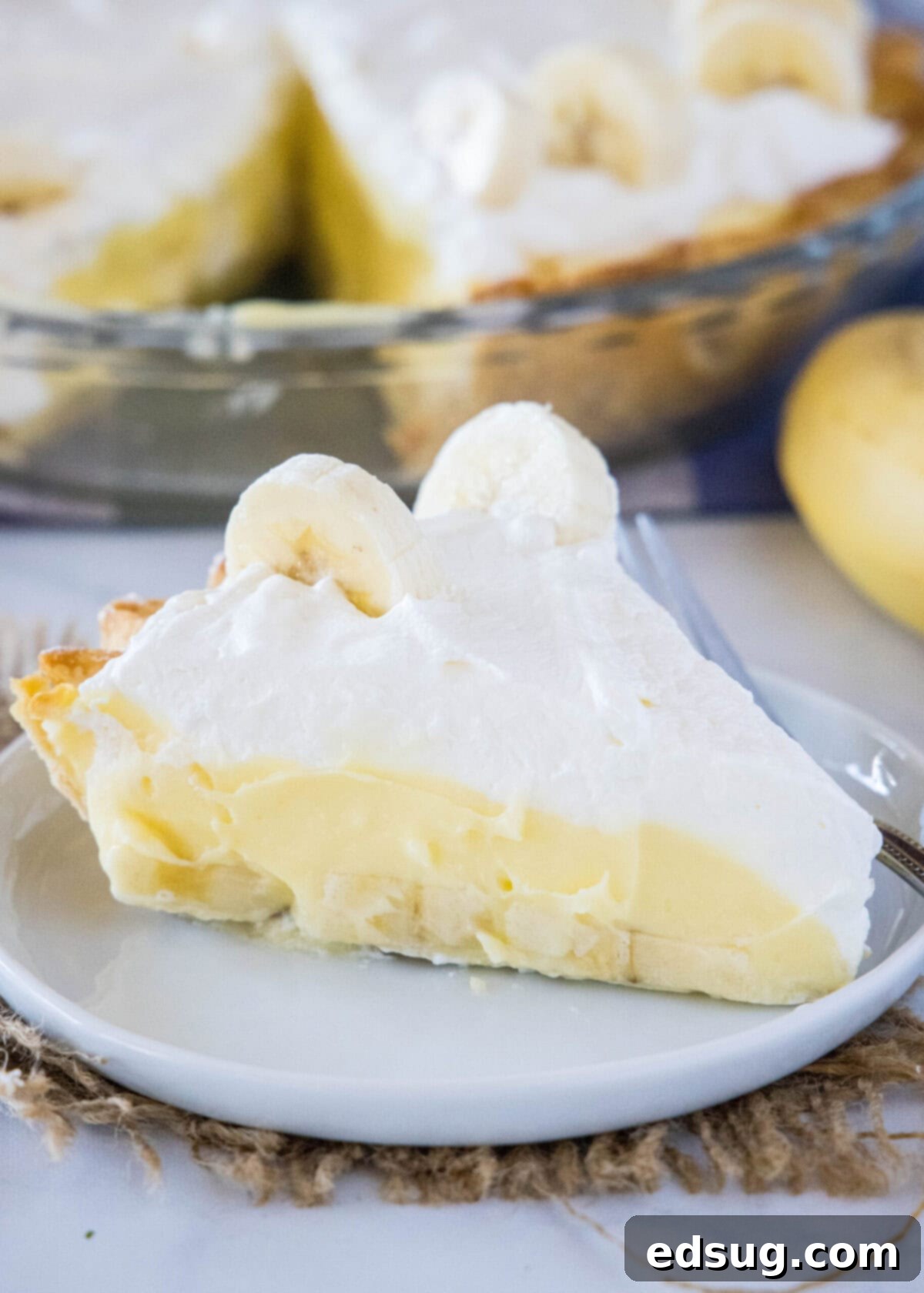 A generous slice of homemade banana cream pie served on a white plate, showcasing its layers of golden crust, creamy banana custard, and fluffy whipped cream, topped with fresh banana slices.